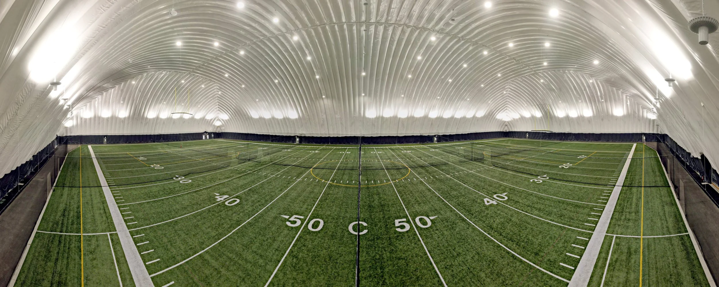 Indoor sports training facility with artificial turf field, white lines, and overhead lighting in a large, enclosed dome
