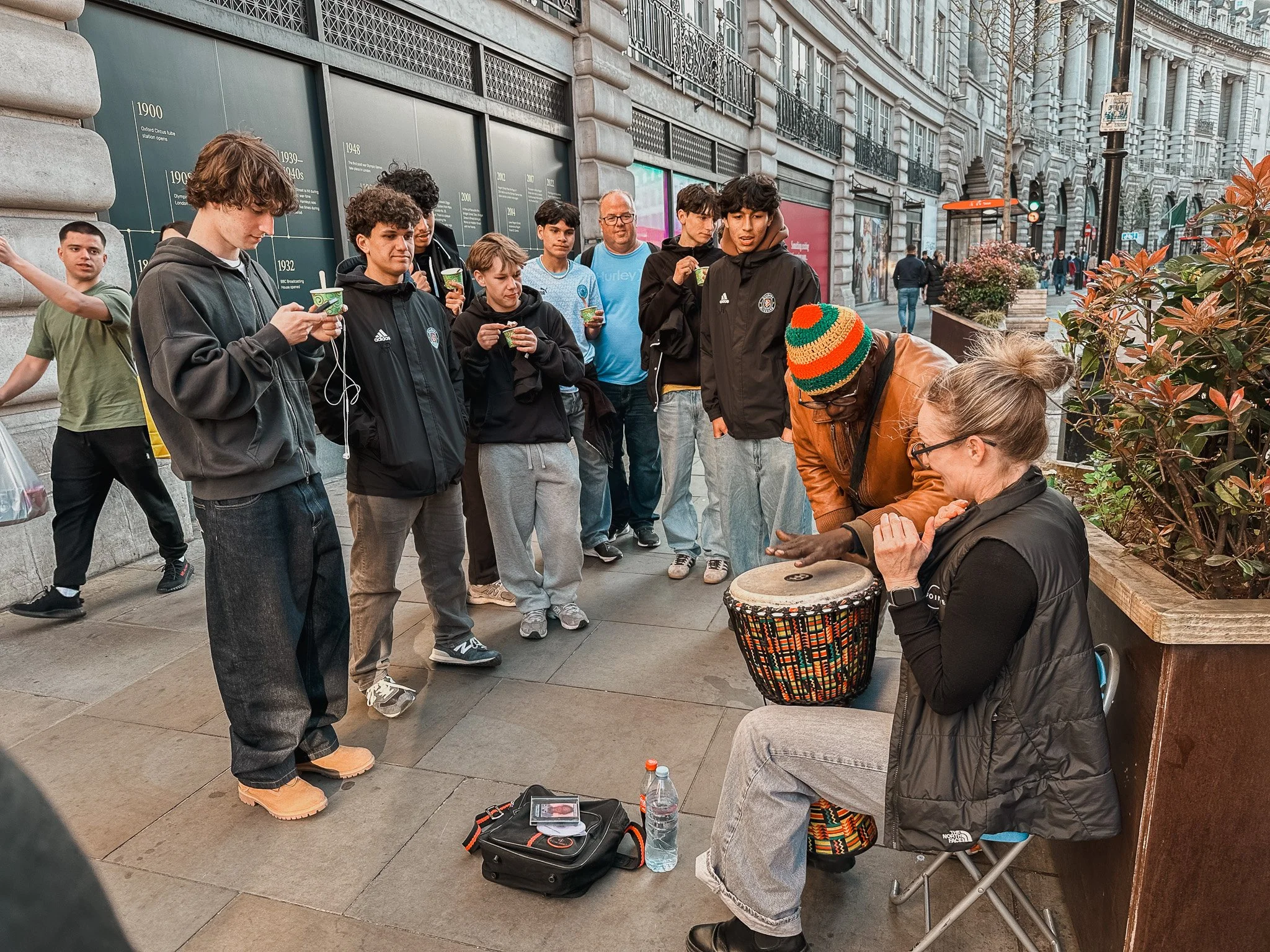 A woman sits on a folding chair on a city sidewalk, playing a drum for a group of people. A man with a colorful hat and gloves watches and appears to join in. Several people are lined up in front, listening, some holding cups. The scene is in an urba
