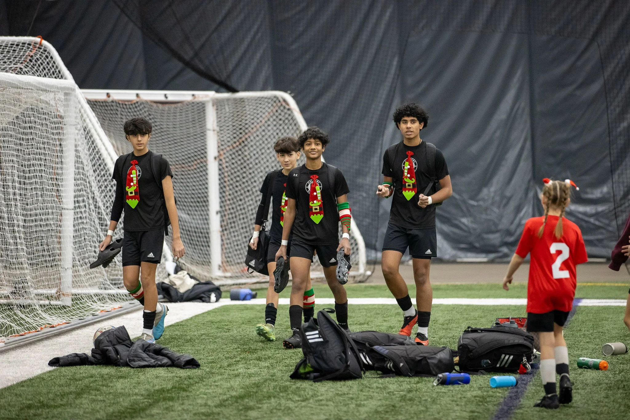 A group of young soccer players walking on an indoor soccer field near the goal, wearing black uniforms with Christmas-themed accessories, with individual bags and water bottles on the ground.