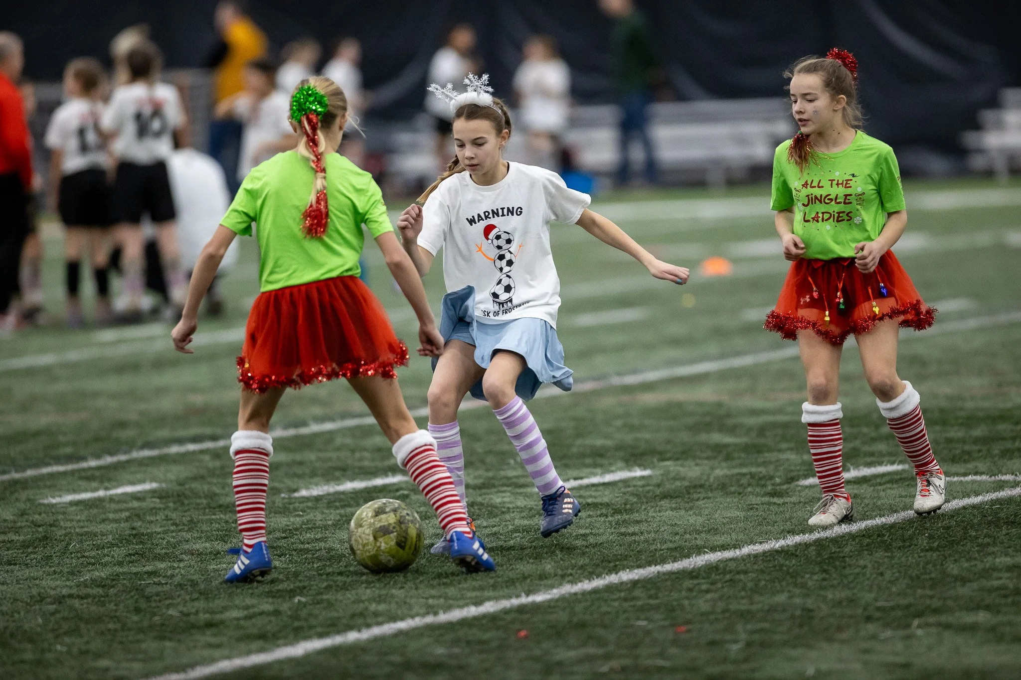 Three young girls playing soccer indoors, wearing festive holiday costumes, with two wearing bright green shirts and red tutus, and one with a white shirt featuring a snowman, on a green turf field, with other kids and spectators in the background.