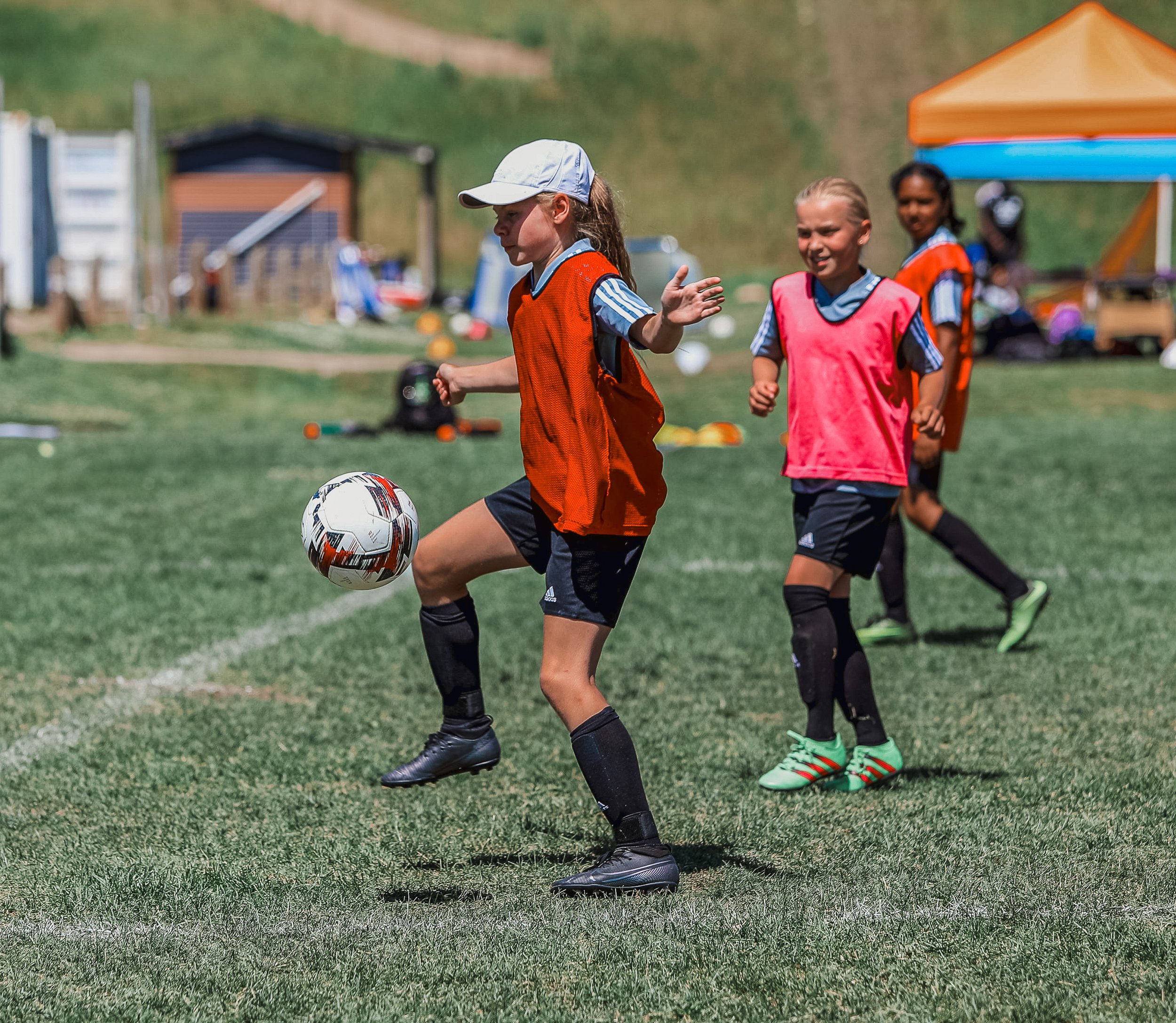 Young girls playing soccer on a grassy field, wearing sports uniforms and training bibs, with a goalpost and outdoor equipment in the background.
