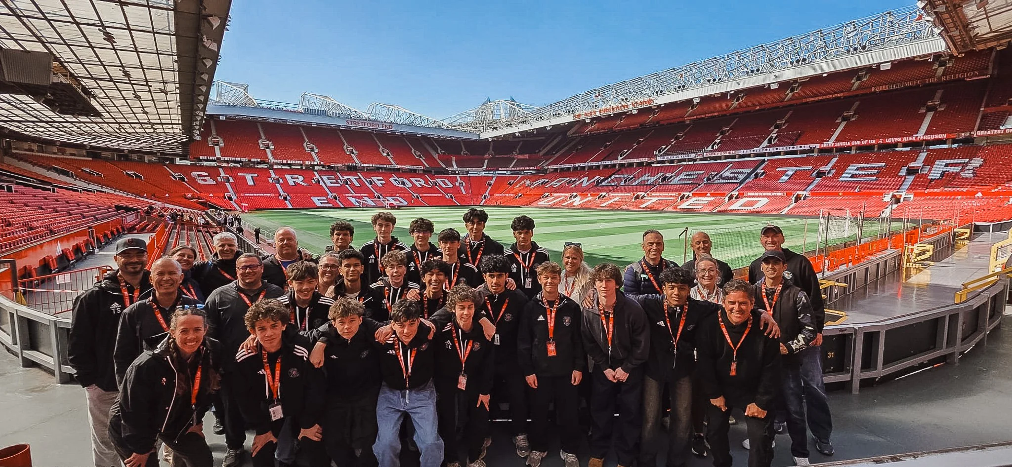 Group of people, including children and adults, standing inside a stadium with red seats and a green field, under a clear blue sky.