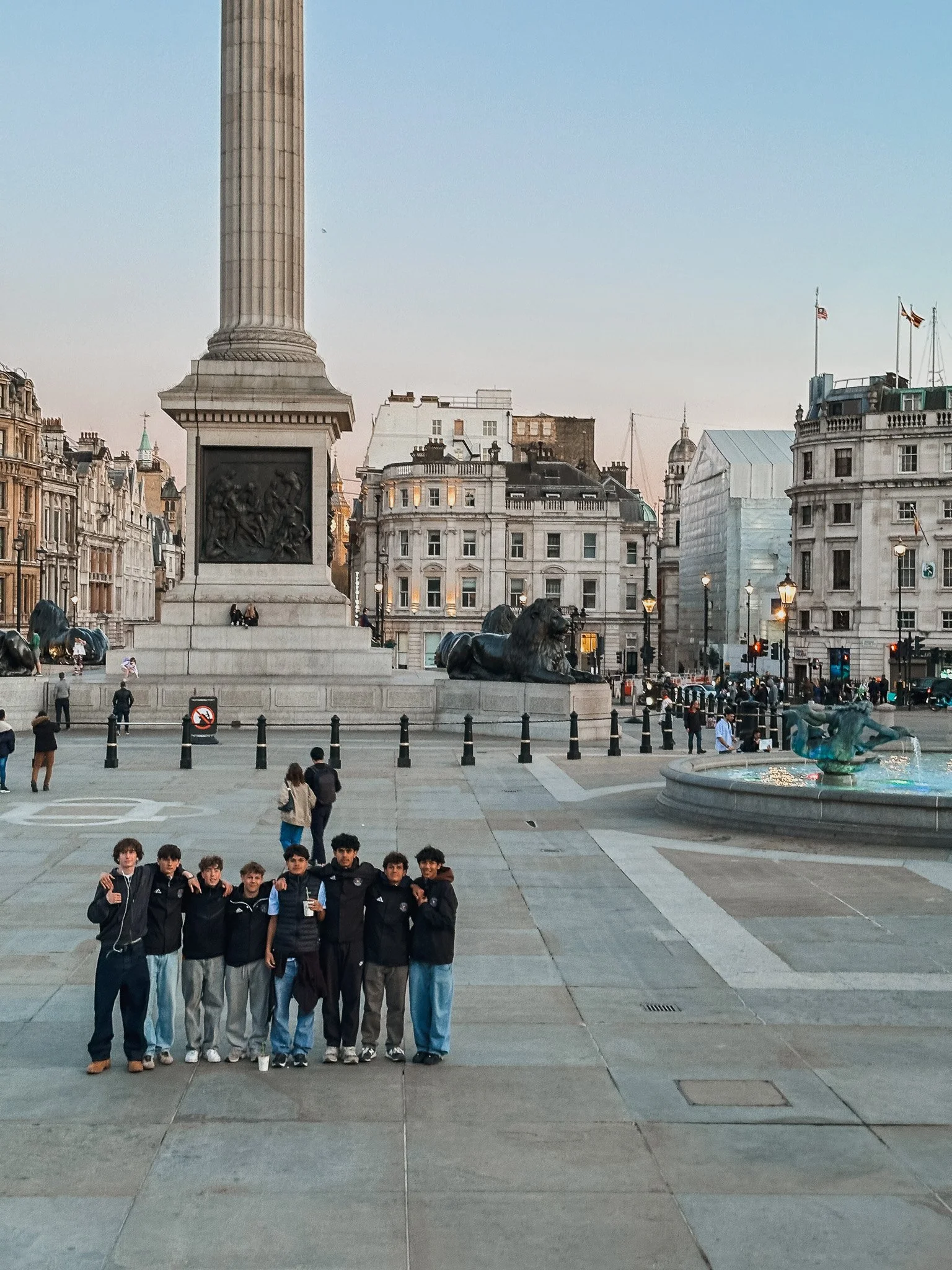 A group of young men standing together in a city square with historical buildings, statues, and a fountain behind them, likely in London.