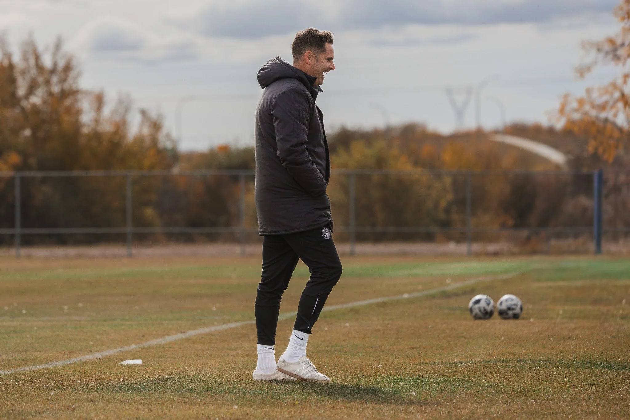 A man dressed in black athletic clothing standing on a soccer field near two soccer balls, smiling with hands in pockets, during daytime with cloudy sky.