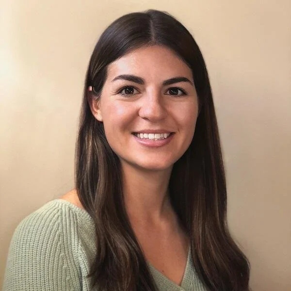 A woman with straight dark brown hair and a light green sweater, smiling against a beige wall.