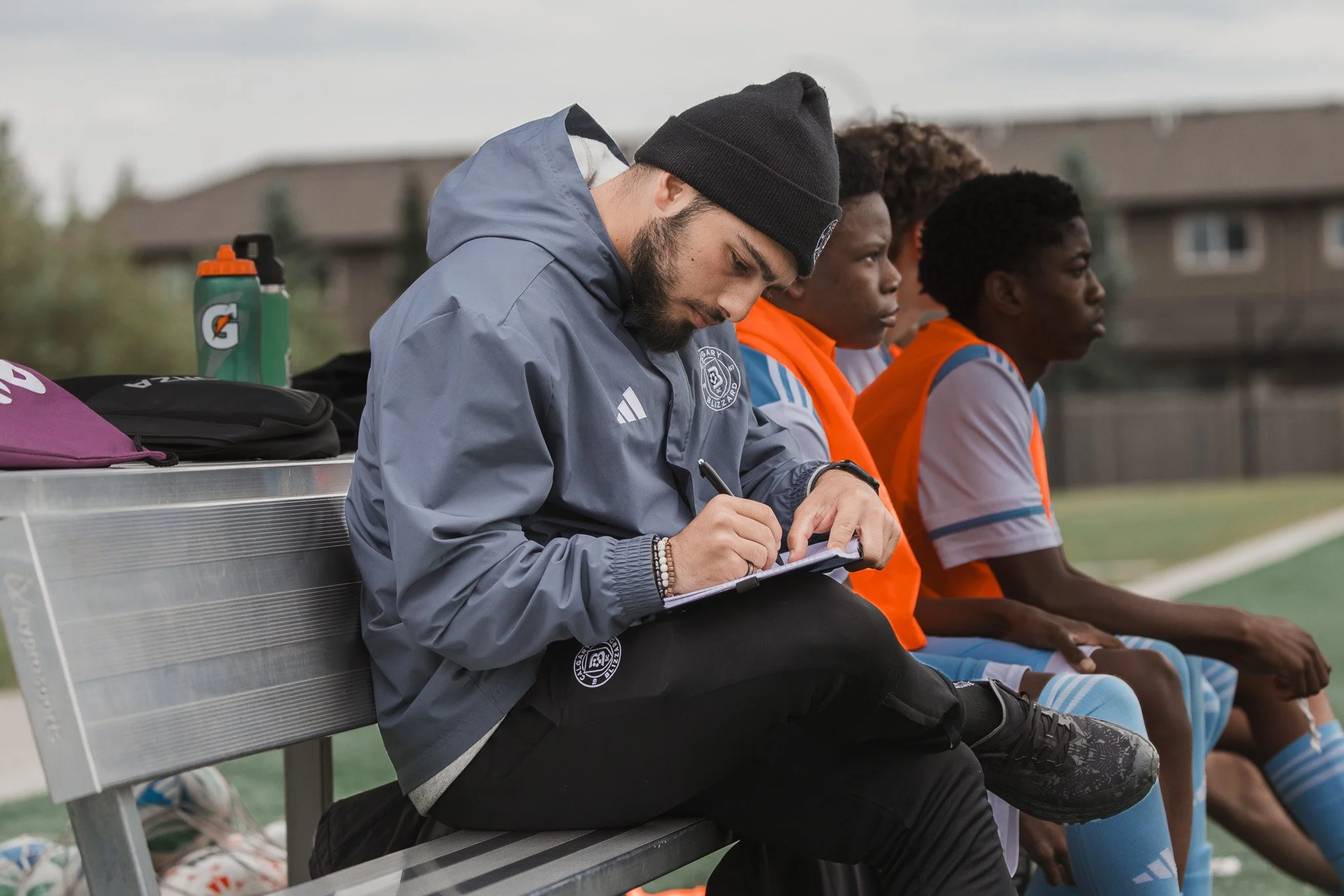 A coach sitting on a bench, writing in a notebook during a soccer game, with players sitting beside him on the bench.