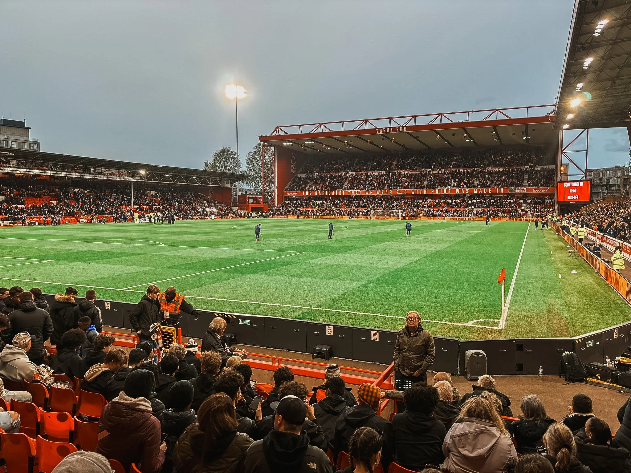 A football stadium filled with spectators before a game, with players warming up on the field, and a cloudy sky overhead.