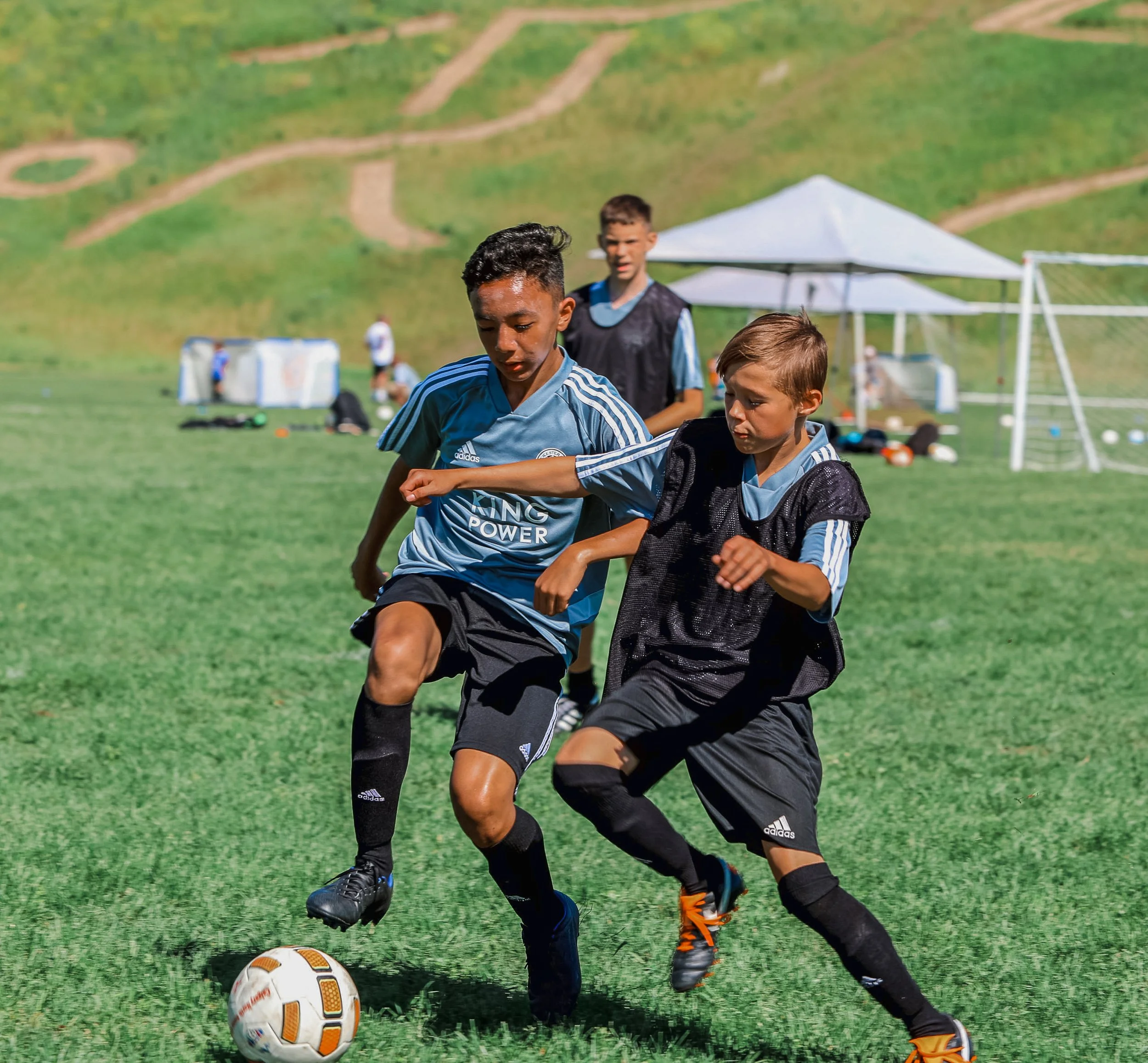 Young boys playing soccer on a grassy field during practice, with a tent and soccer goals in the background.