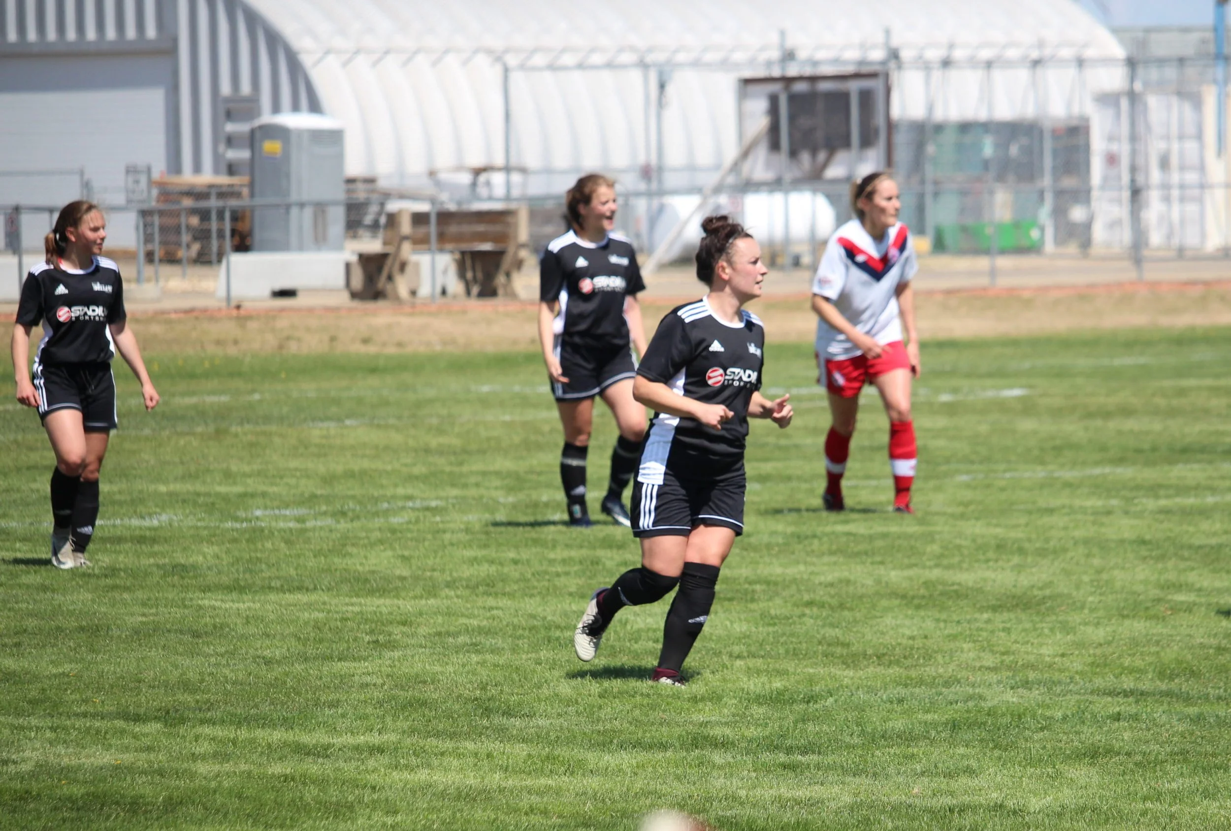 Female soccer players on a green field during a game. Four players are visible, two in black uniforms, one in a white and red uniform. The players are running and appear to be enjoying the game. There is a fenced area and some structures in the background.