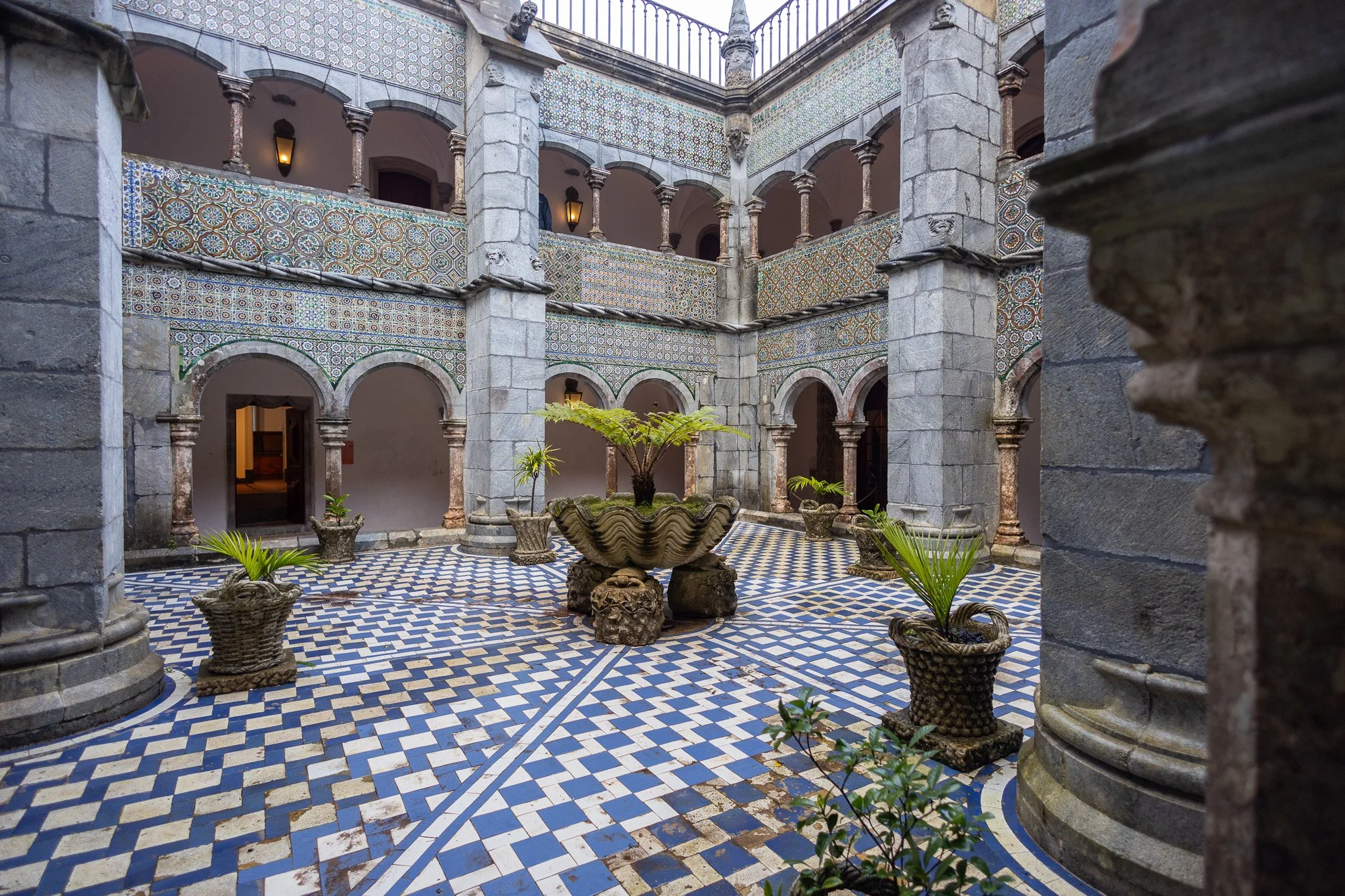 Interior courtyard with stone columns and arches, featuring colorful geometric tile patterns on the walls and floor, potted plants in ornate containers, and a central decorative fountain with lush green plants.