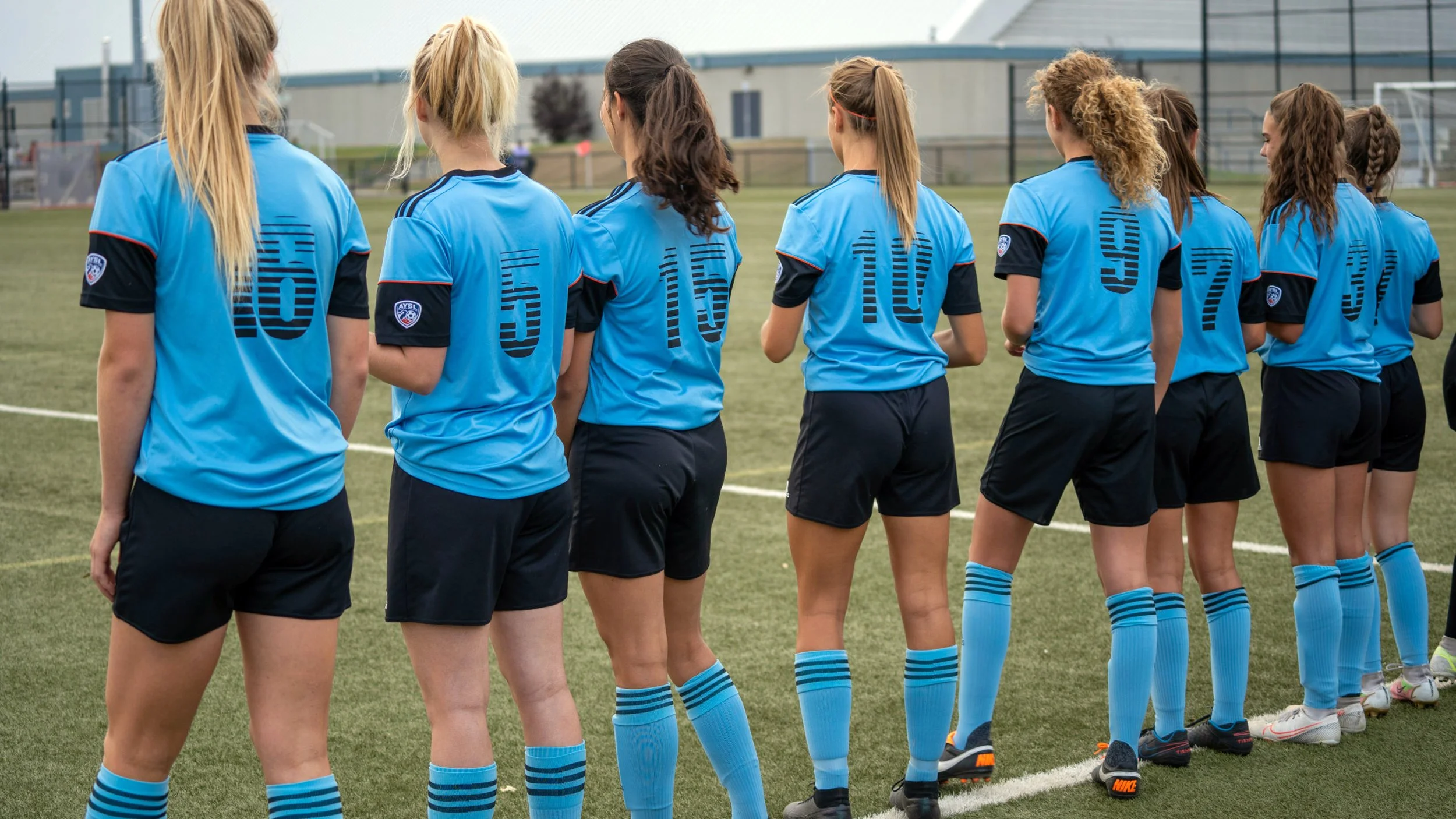 A group of female soccer players standing in a line on the field, wearing blue jerseys and black shorts, with some having socks pulled up and wearing soccer shoes.