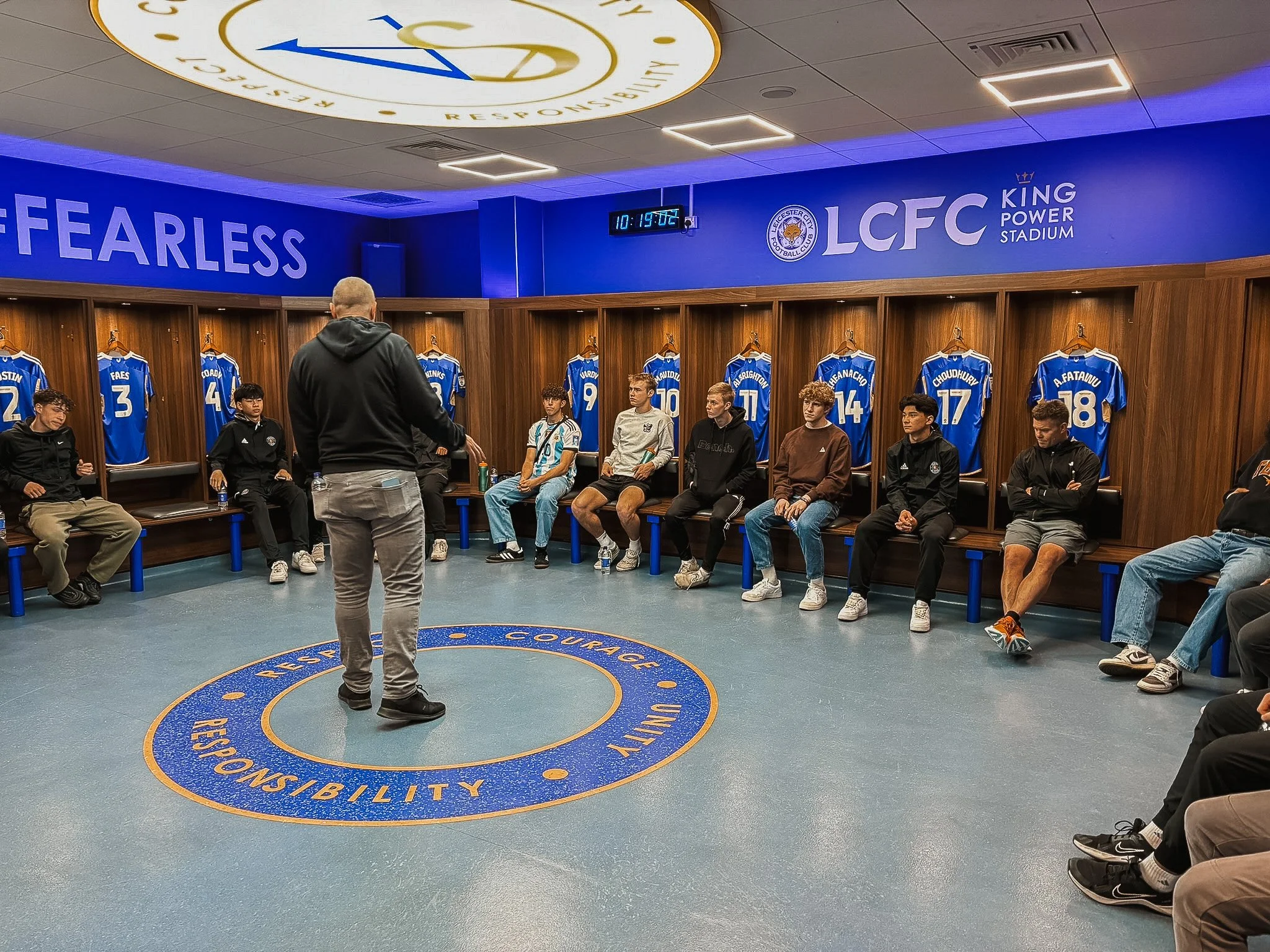A group of young men sitting on chairs in a locker room with blue Leicester City football jerseys hanging behind them, listening to a coach or speaker standing in front of them, with the Leicester City logo visible on the wall and a circular motto on