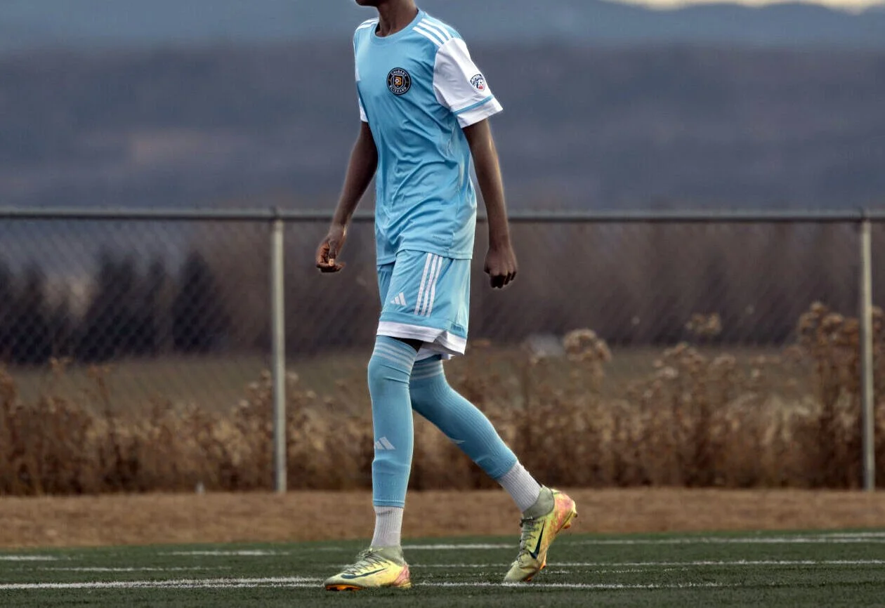 A soccer player standing on a field wearing a light blue uniform, yellow cleats, and long light blue socks.