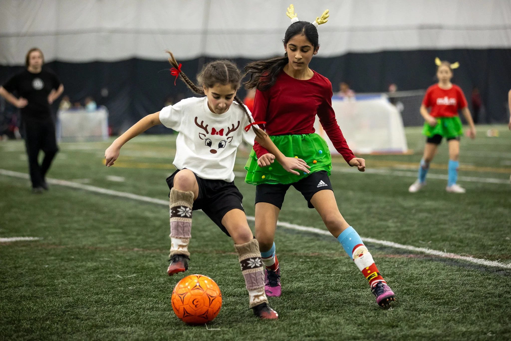 Two young girls playing soccer indoors, competing for the orange soccer ball. One girl has braided hair with a red bow and Santa-themed socks, and the other girl has long dark hair with reindeer antlers headband and colorful socks. Other children and