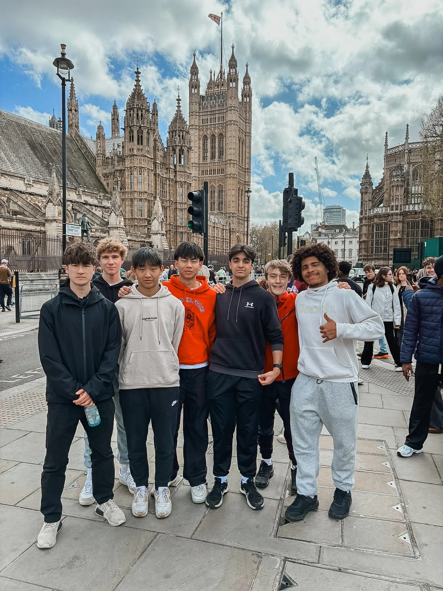 A group of seven young men posing in front of the Palace of Westminster in London, with the iconic gothic-style buildings and a partly cloudy sky in the background.