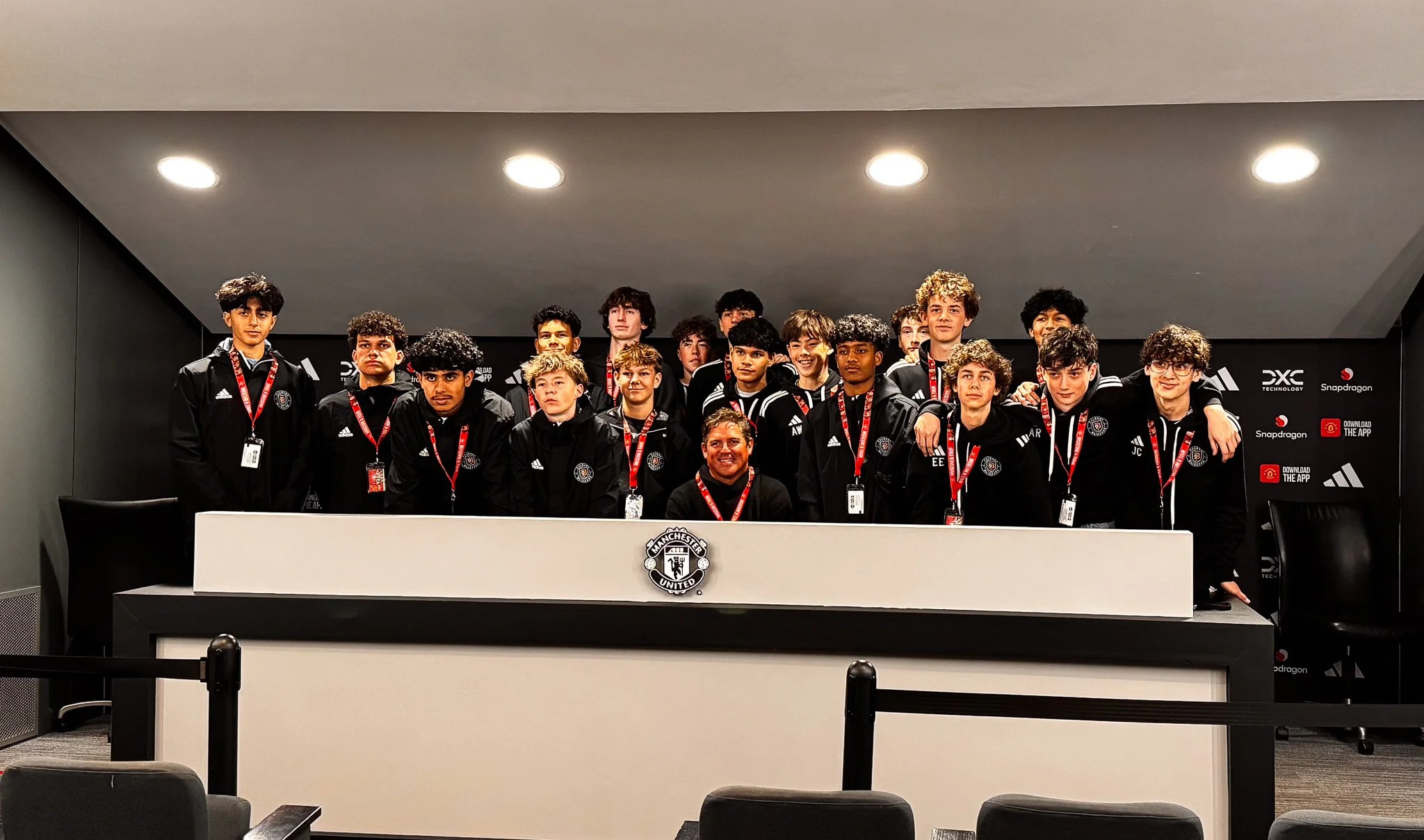 Group of young men and a coach posing for a photo at a press conference or media event, in front of a black backdrop with sponsor logos, wearing black jackets with Manchester United logos and event badges, on a stage with a white table and Manchester