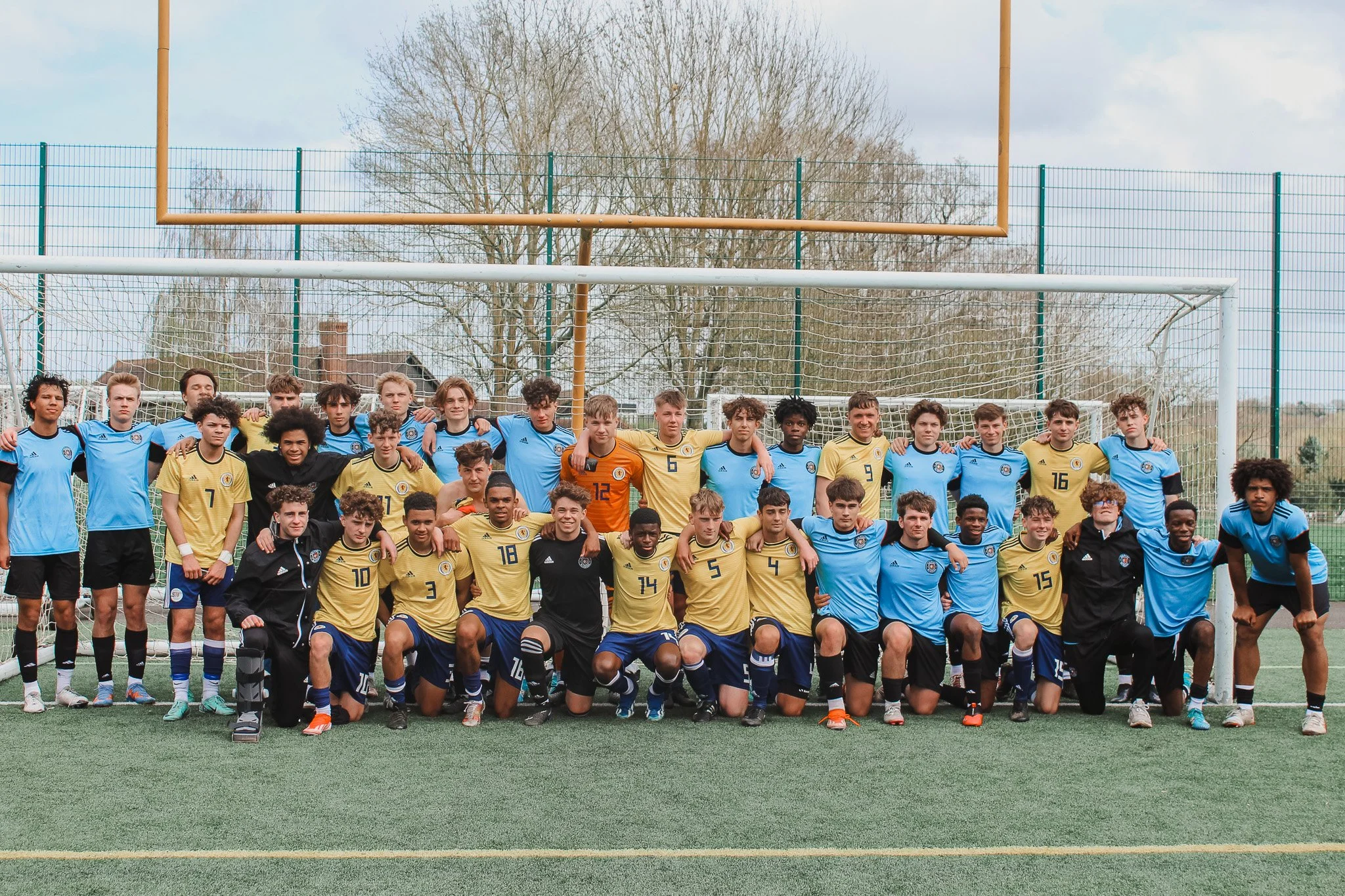 Group of young soccer players and coaches posing for a team photo on a soccer field, with goalposts and a fence in the background.