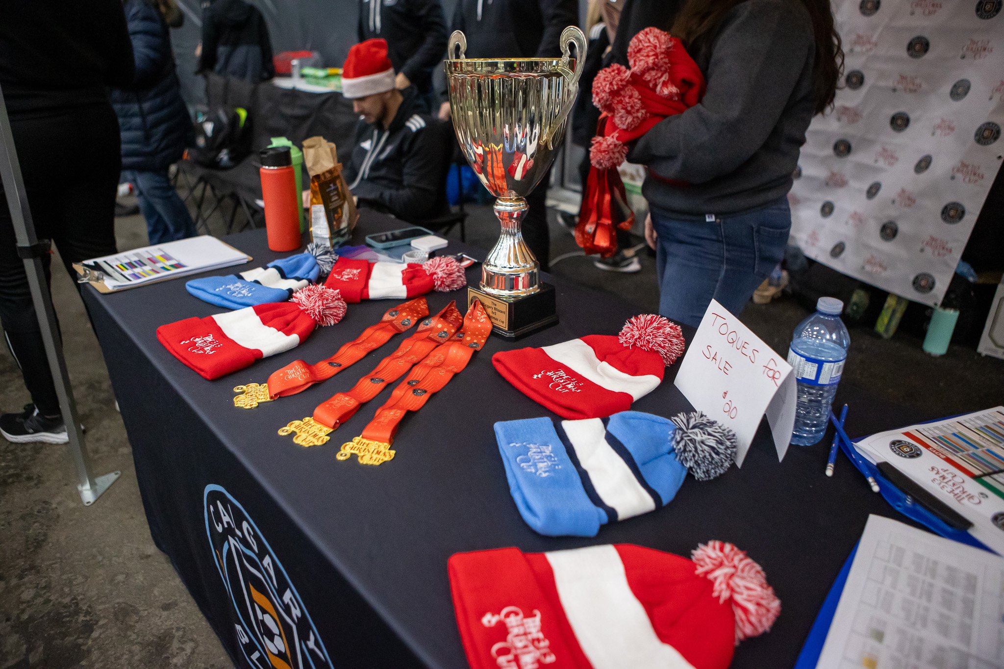 A table with medals, Christmas hats, and a trophy, at a holiday event or race. People are in the background, some wearing Santa hats, and there is a sign indicating an item for sale.