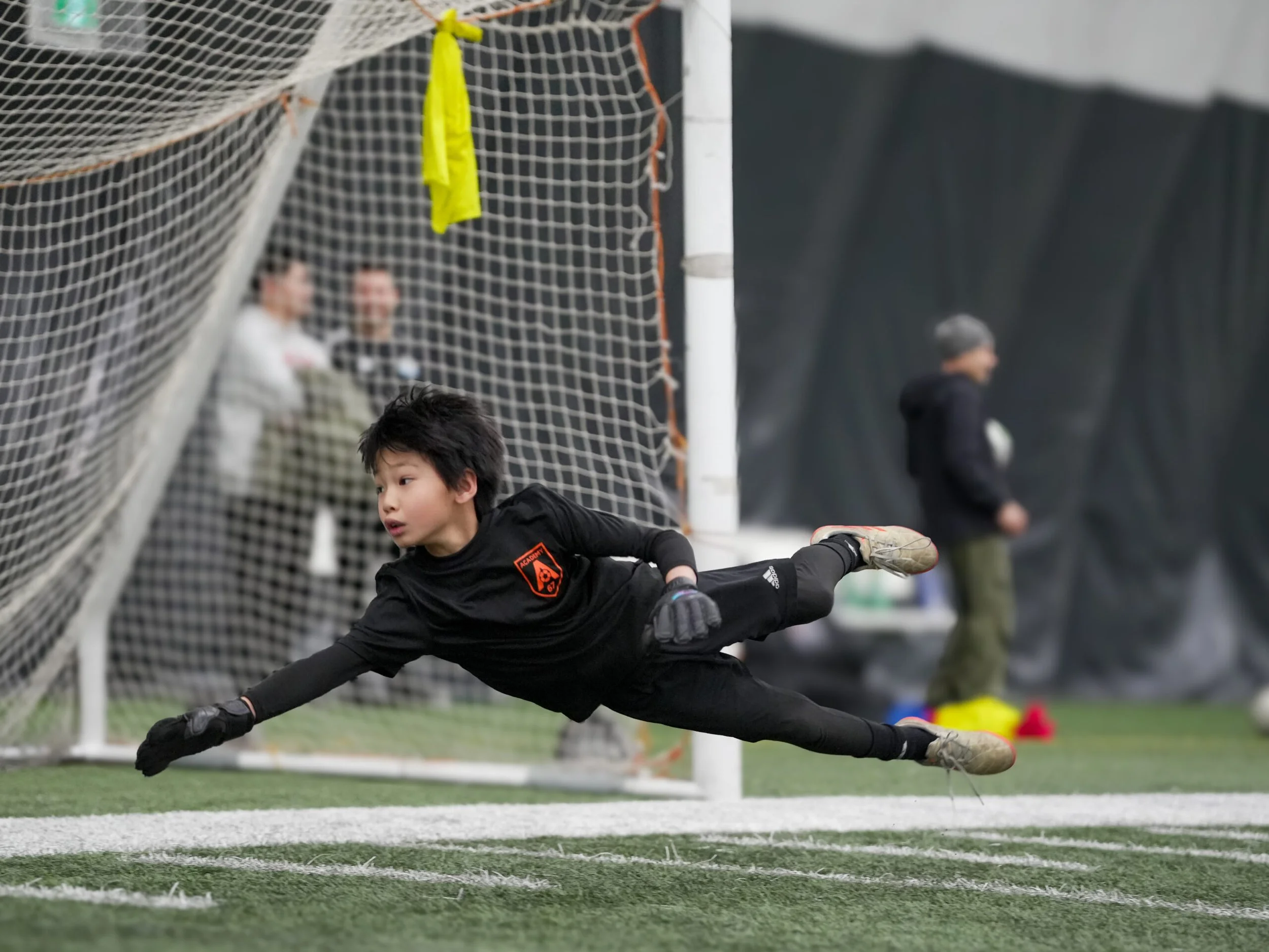 A young boy soccer goalie diving to save a ball during practice or game inside an indoor sports facility.