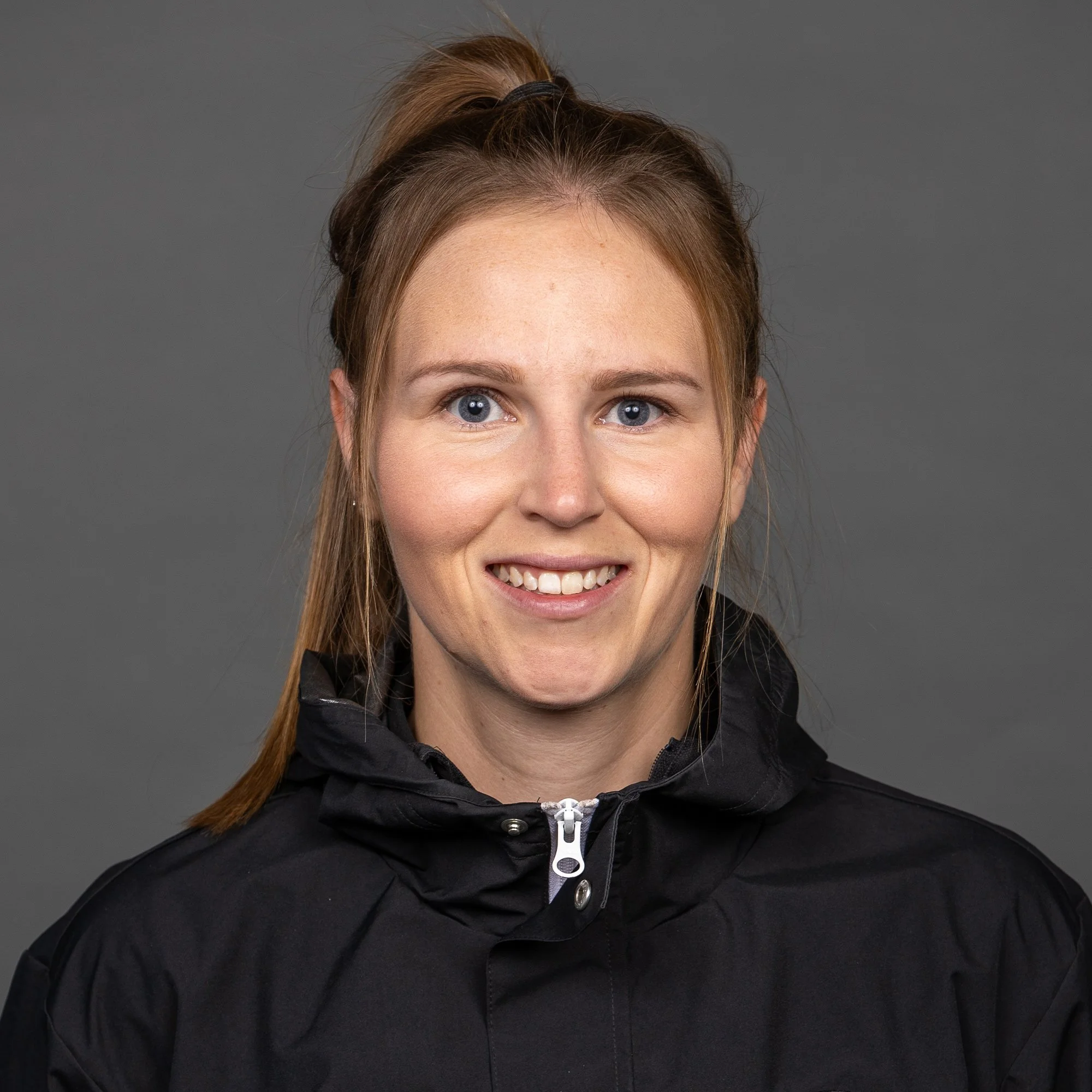 A young woman with blonde hair and blue eyes standing outdoors at night, wearing a black Adidas sports jacket with a logo on the chest.