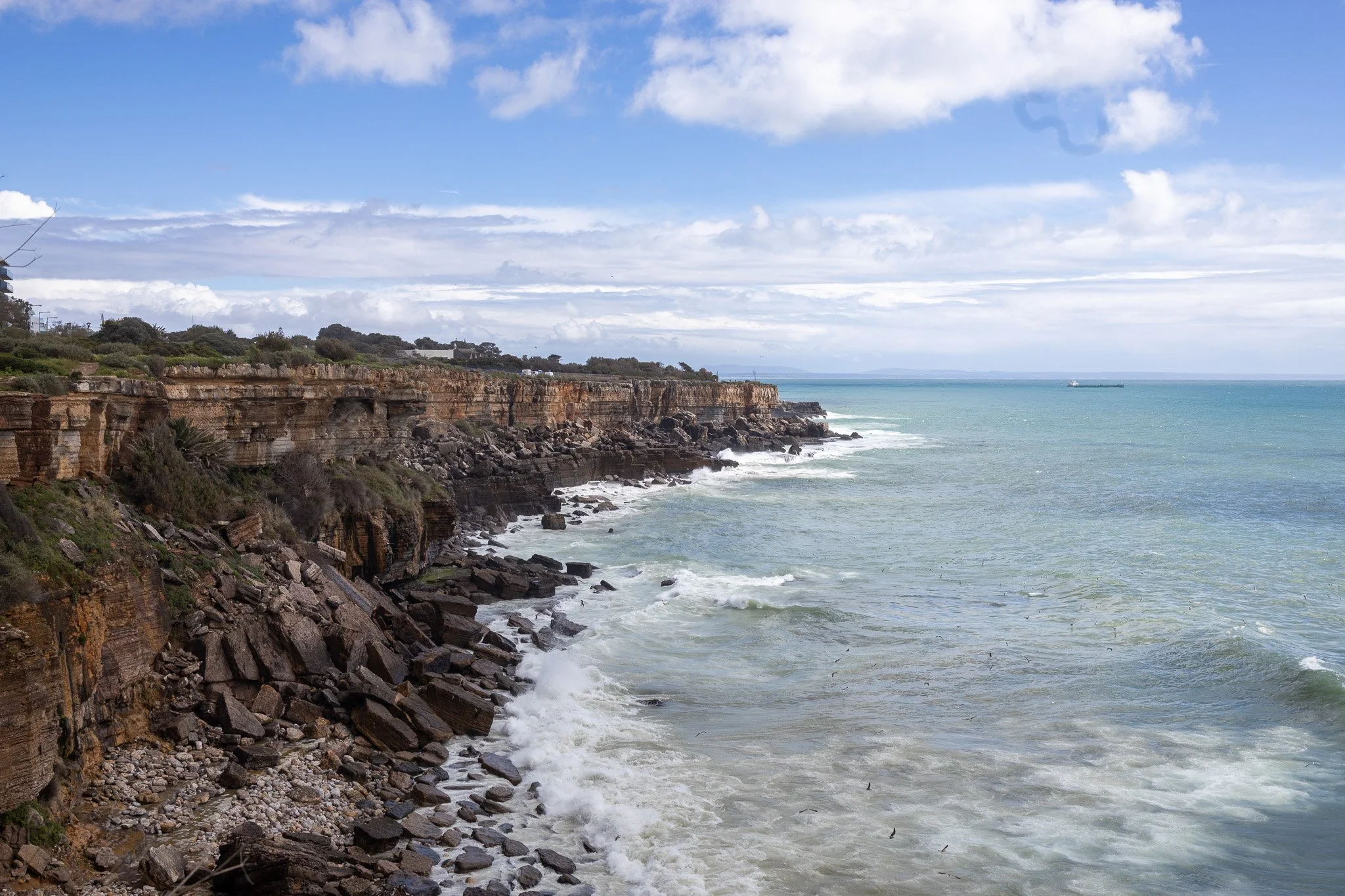 Coastal cliffs with vegetation on top, rocky shoreline below, and ocean waves under a partly cloudy sky.