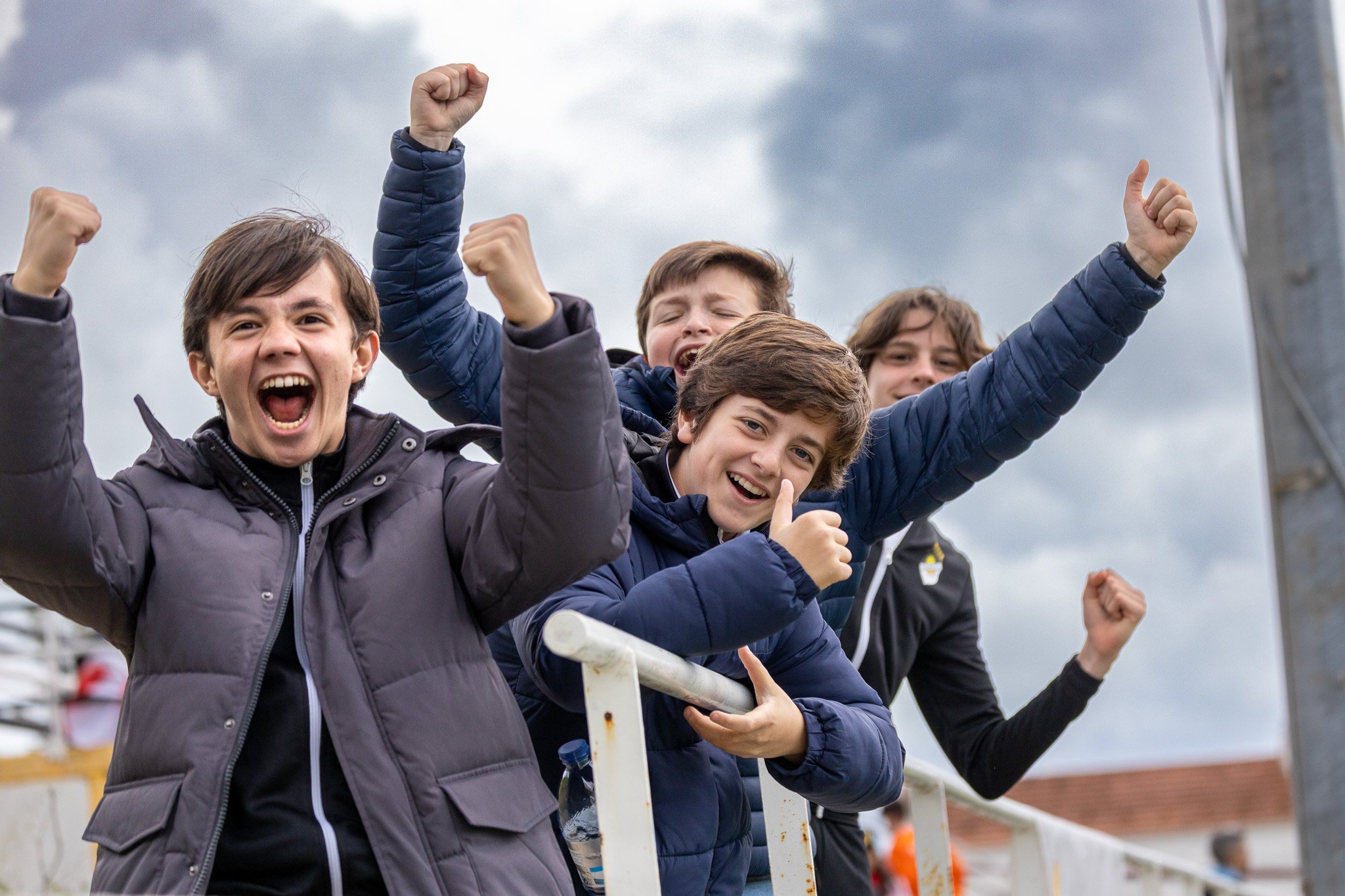 Group of four boys celebrating outdoors, cheering and smiling with fists raised, under cloudy sky.