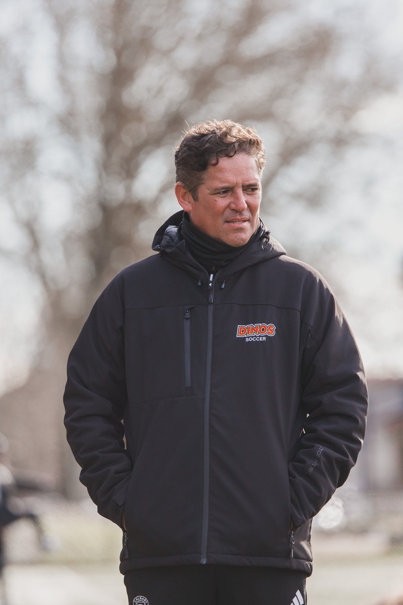 A man wearing a black jacket with the logo 'DINOS SOCCER' stands outdoors on a cloudy day, looking to the side.