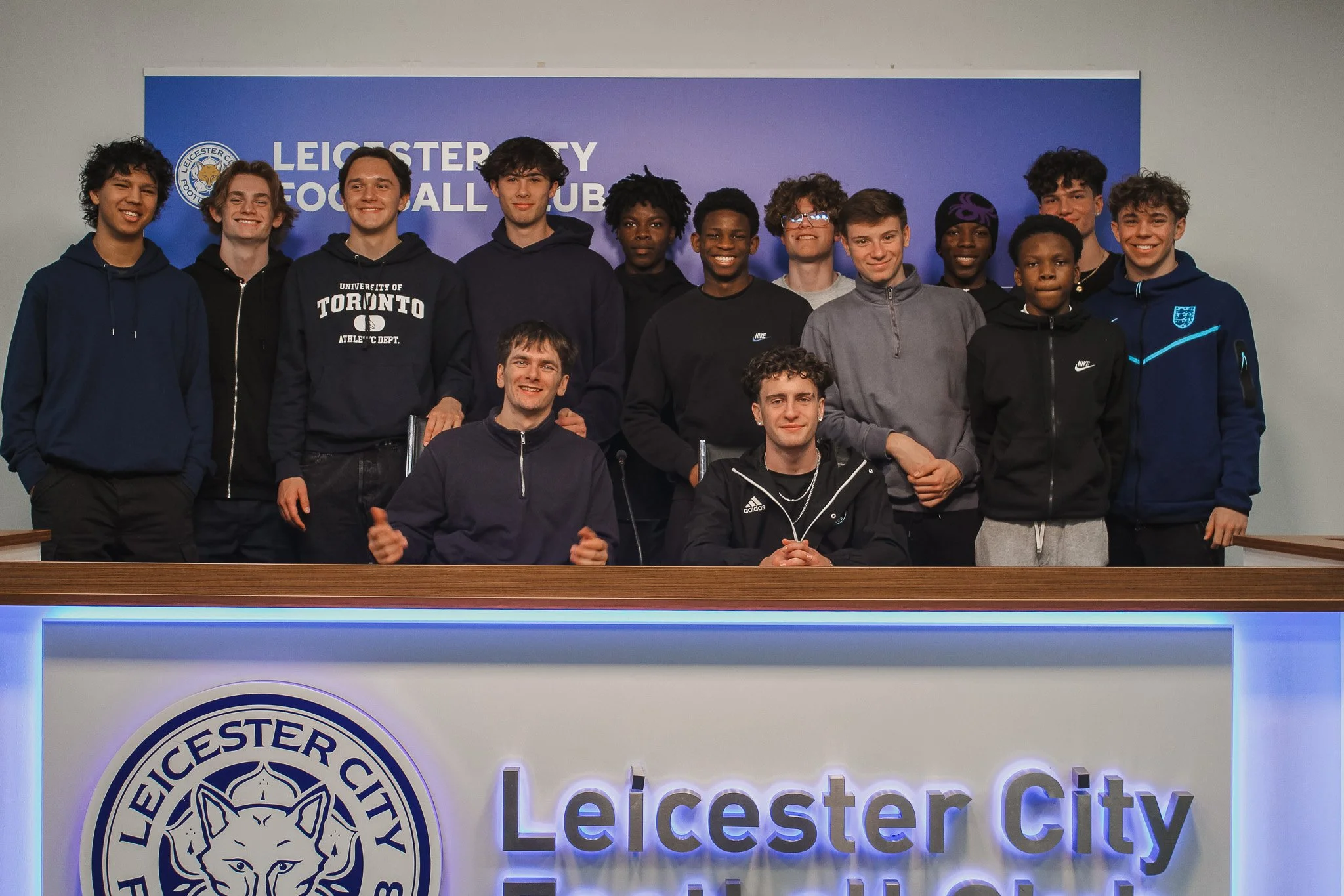 Group of young male athletes and two adults at a press conference or meeting, with a Leicester City Football Club sign in the background.