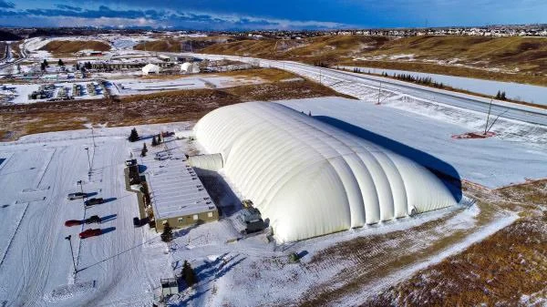 A large white inflatable dome structure in a snowy landscape, with some smaller buildings and parked vehicles nearby.