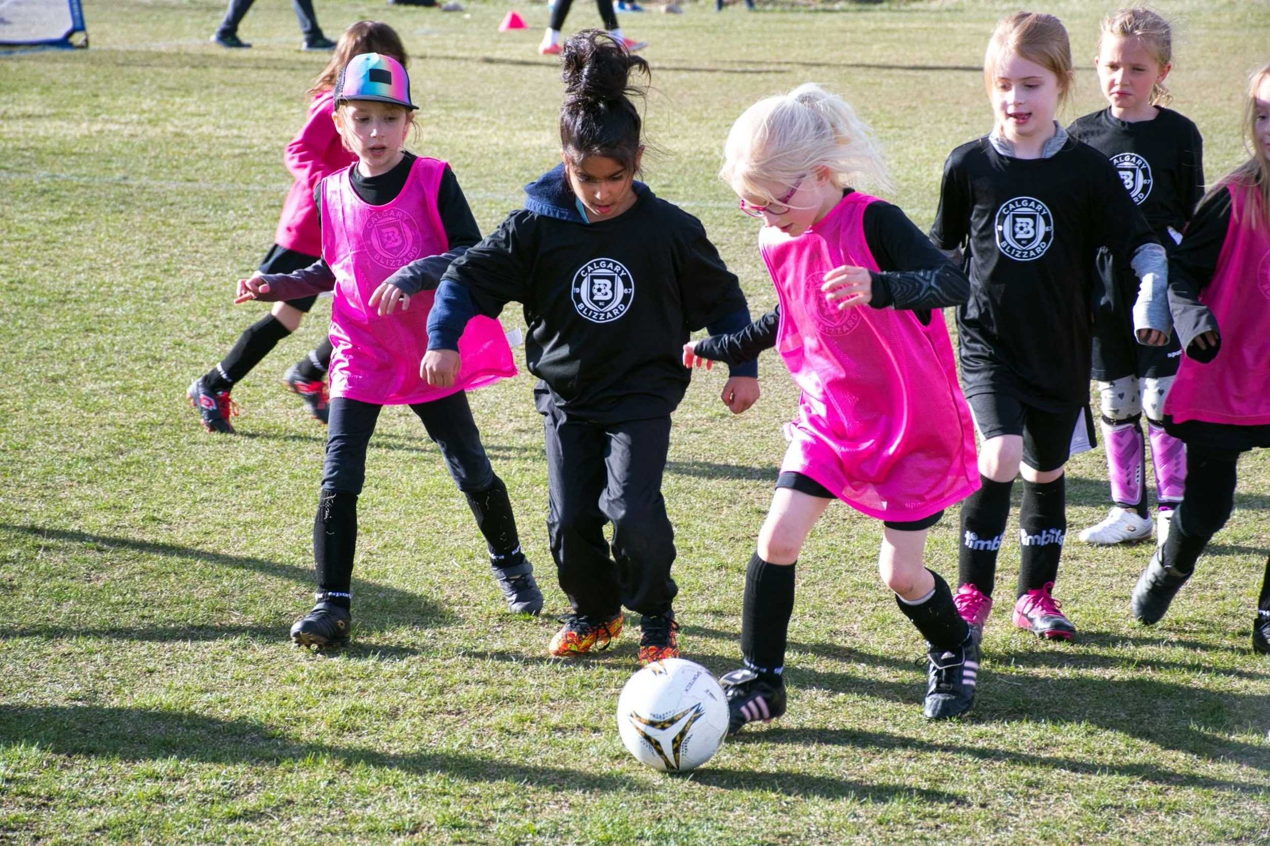 Young girls playing soccer on a grassy field, one girl kicking a ball while others watch and prepare to join.