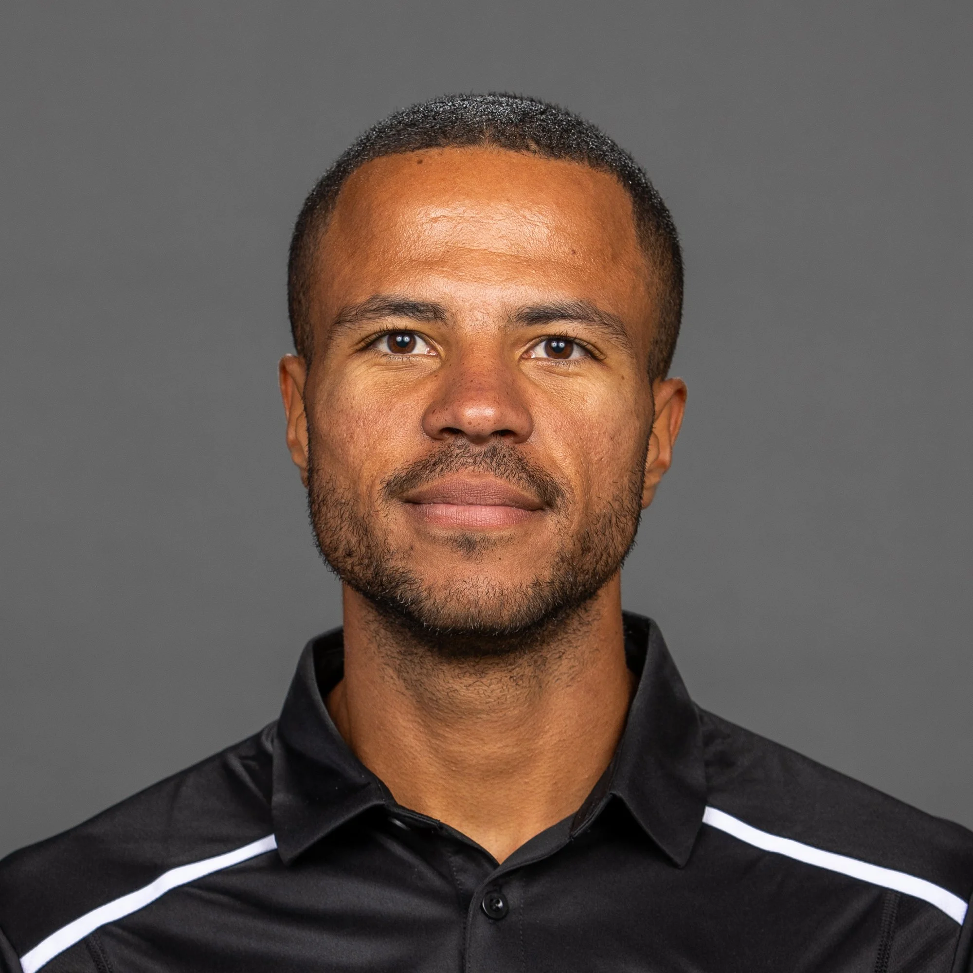 A man with short dark hair and a beard, wearing a black athletic jacket with the adidas logo and a soccer team crest, standing outdoors at night with blurred lights in the background.