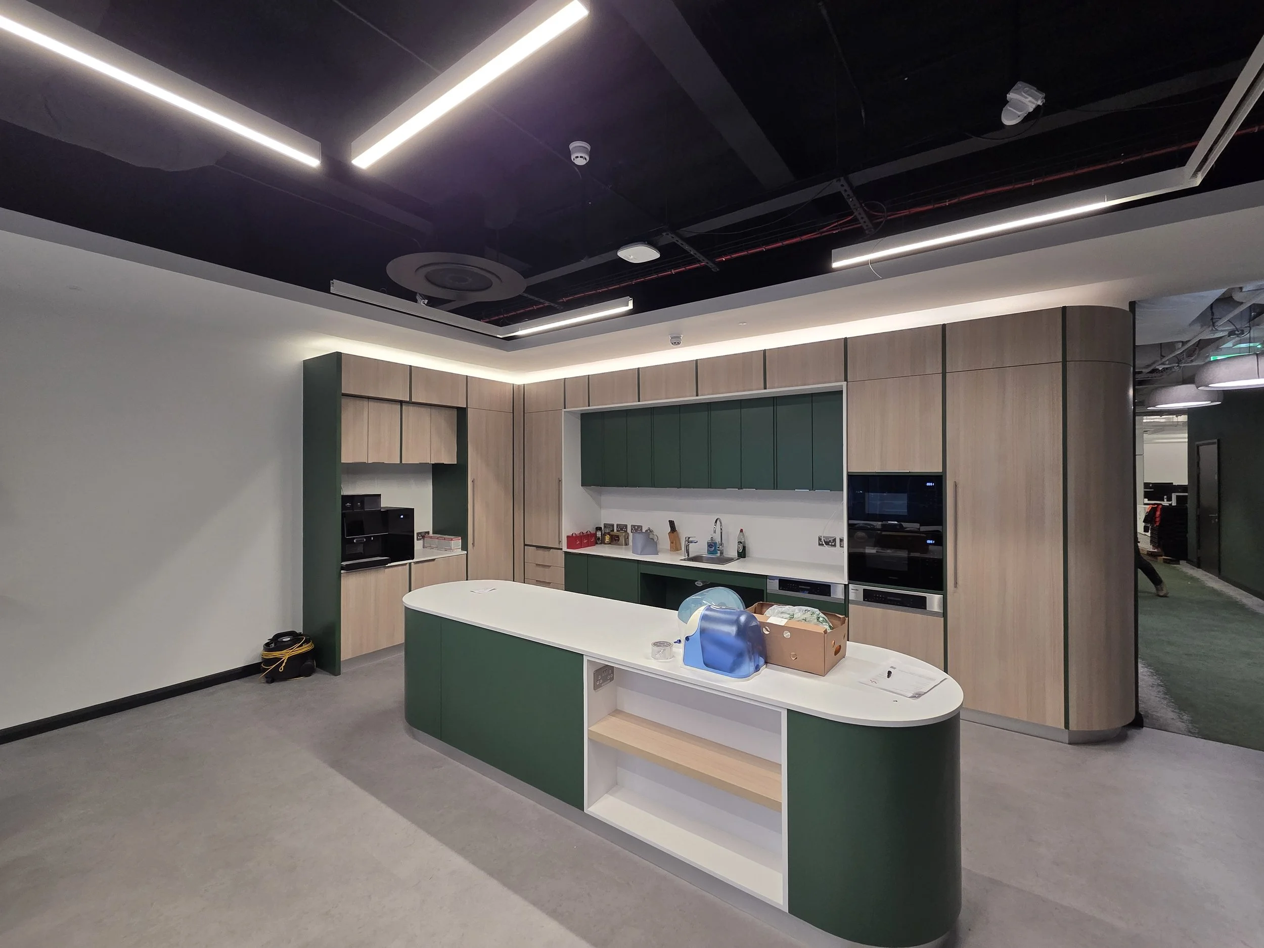 Modern kitchen with feature ceilings and green and wood cabinetry, an island with a white countertop, and built-in appliances, in an office or commercial space.