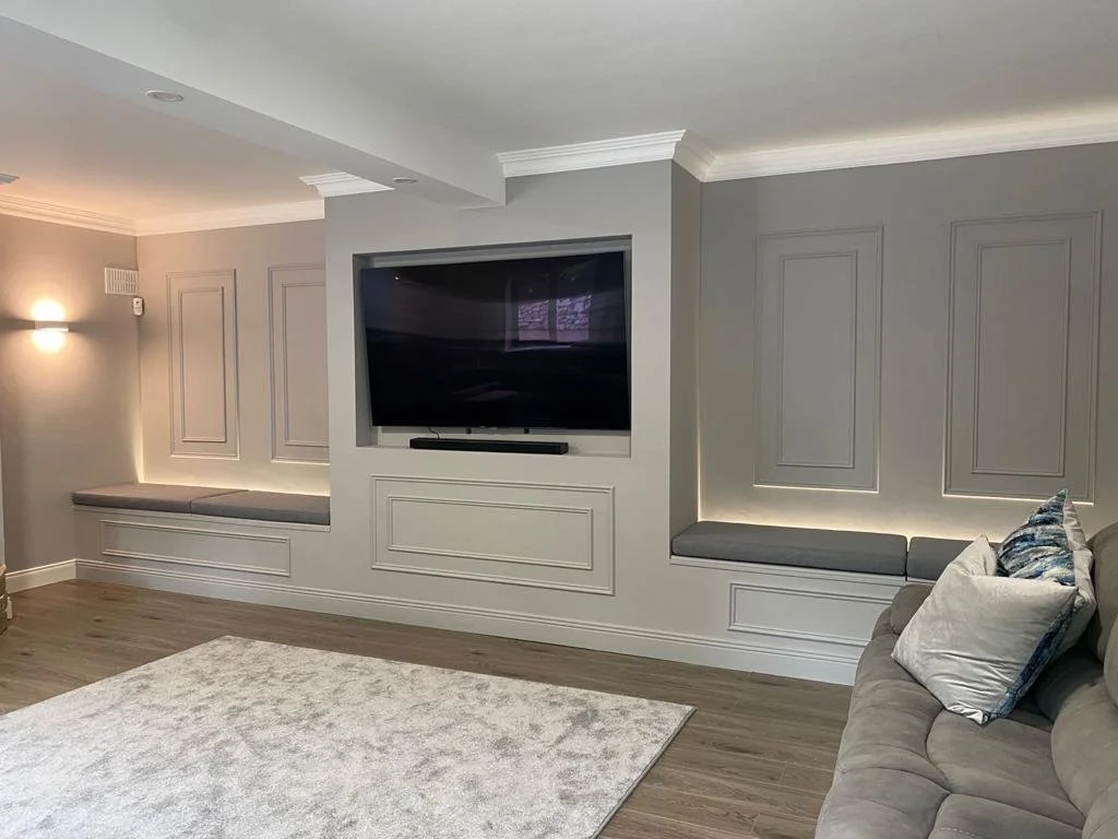 Living room with a built in media wall showing a wall-mounted TV, built-in seating with cushions, gray walls with decorative panel molding, and a plush white area rug.