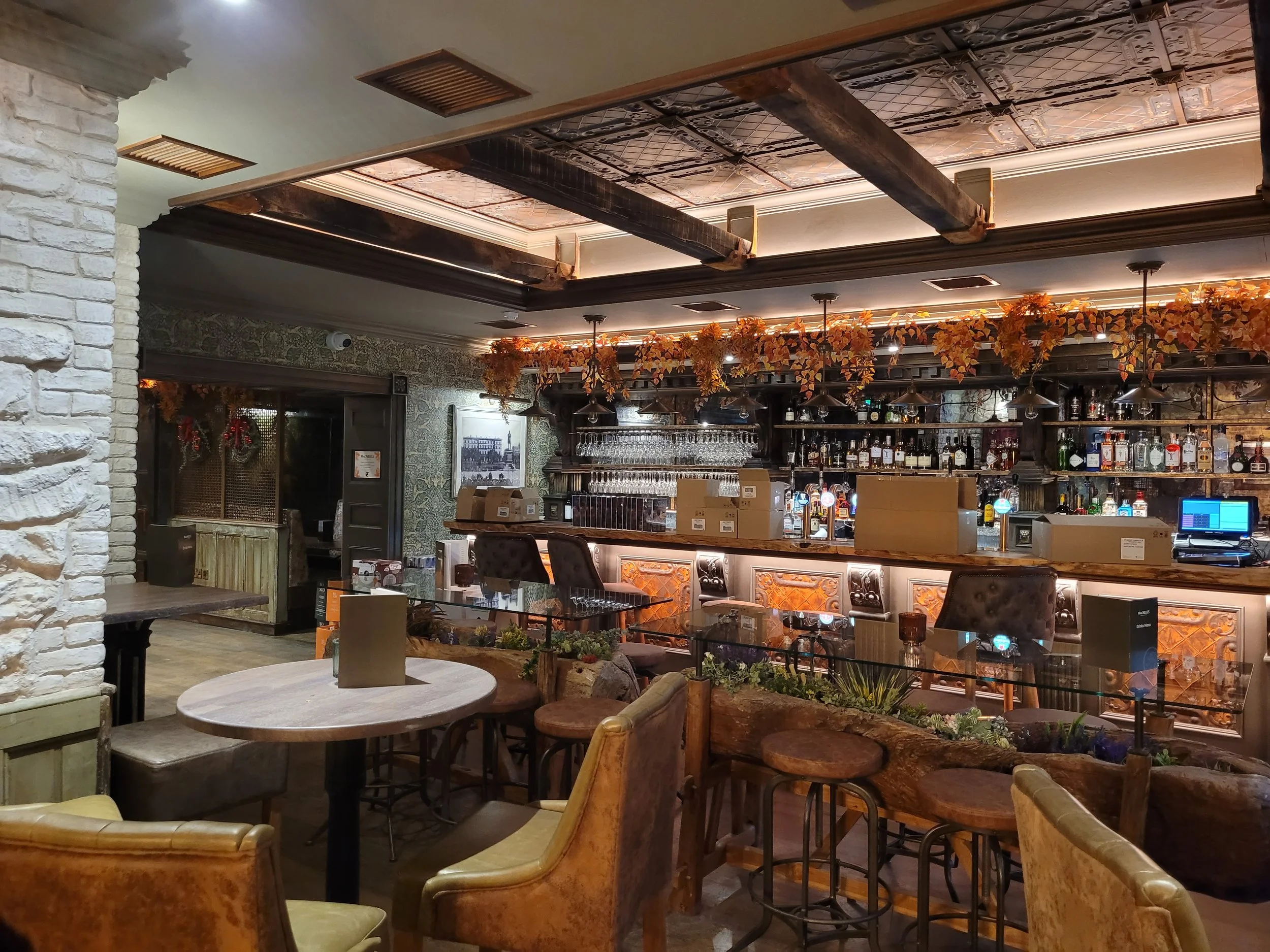 Cozy bar area with a feature ceiling, a wooden bar counter, surrounded by autumn-themed decorations and seating with plush chairs and tables.