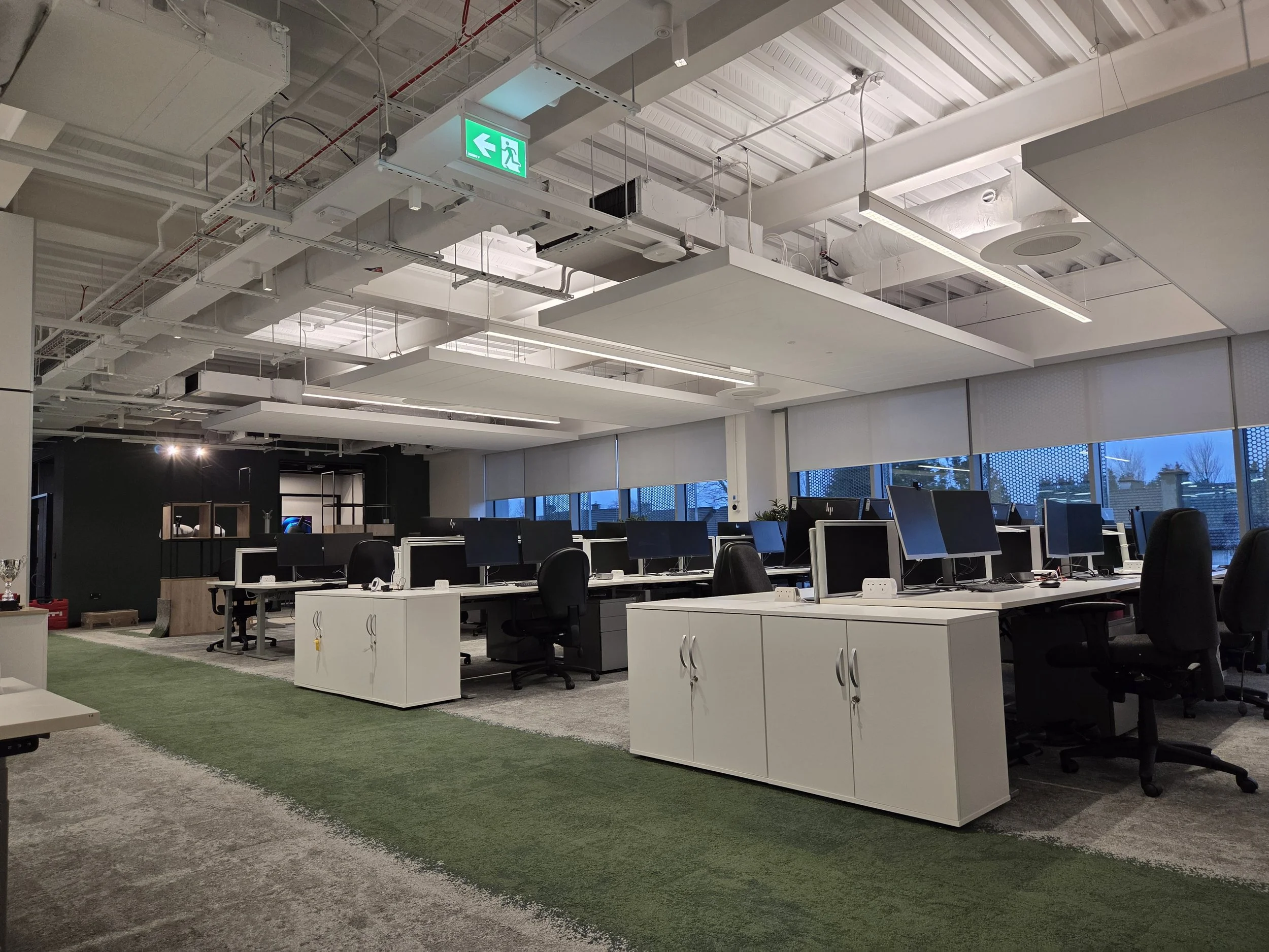 Empty modern office space with multiple desks, computer monitors, and black chairs, overlooking outside trees through large windows, with various styles of ceiling works.