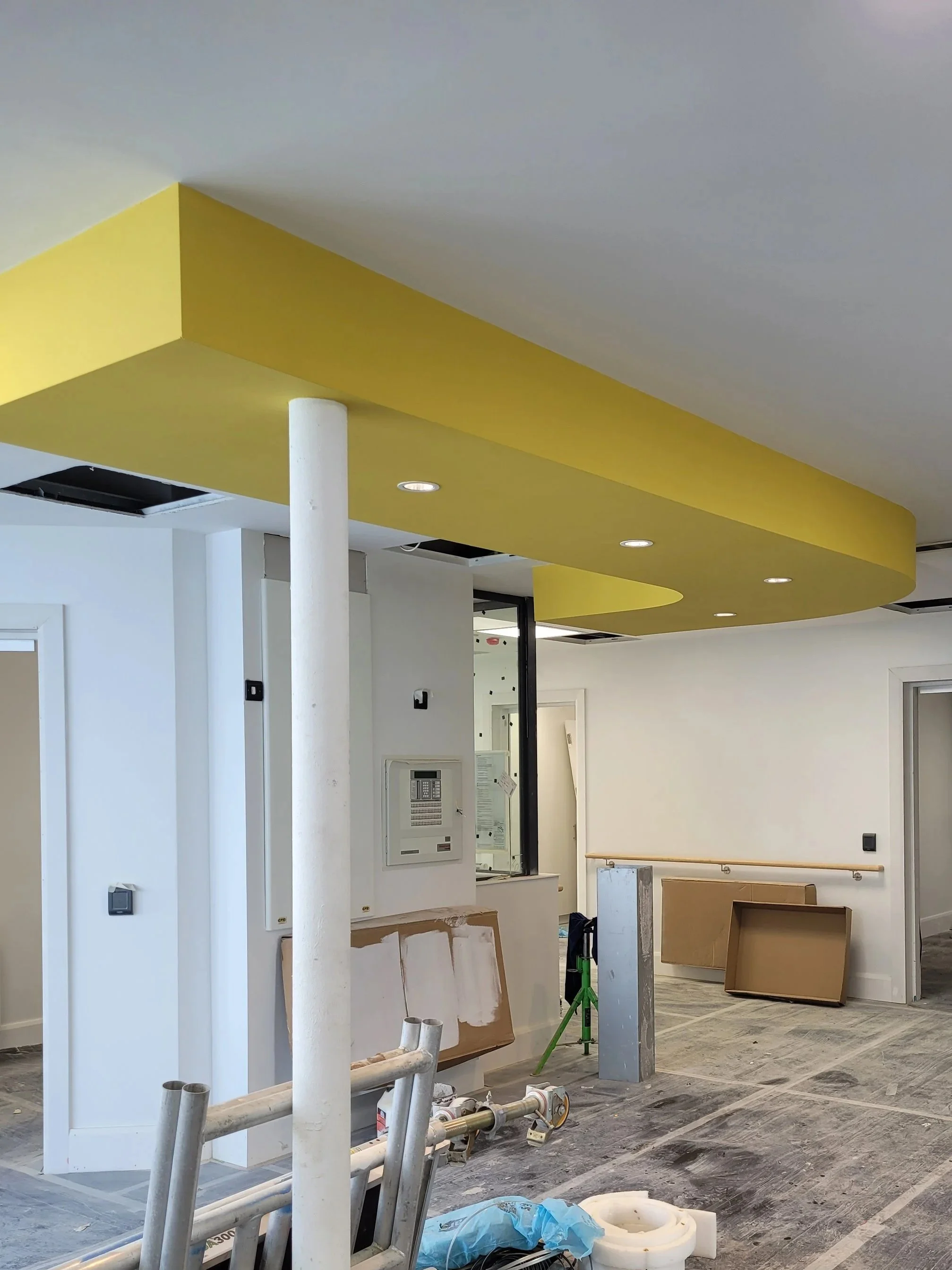 Interior of a building under construction with a yellow ceiling feature, white walls, construction tools, and materials.