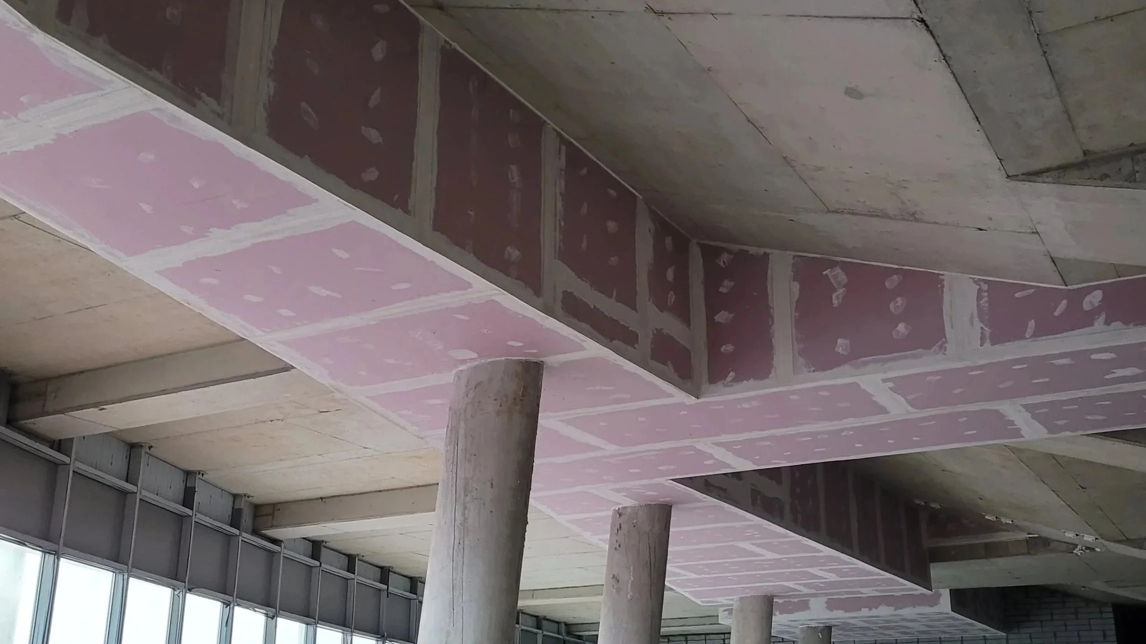 Interior view of a building under construction, showing concrete columns, a partially finished ceiling with pink drywall, and large windows, which highlights fireproofing work.