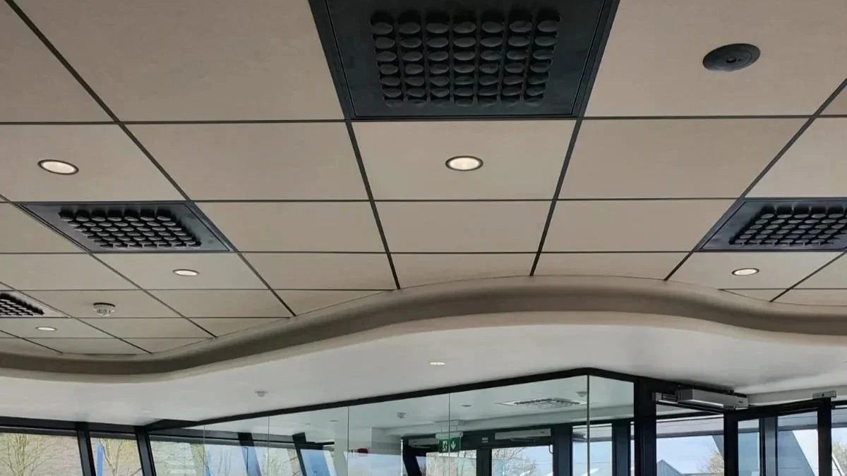 Grid ceiling with square tiles, some with ventilation panels, and a wavy edge border, inside a modern building with large glass windows.