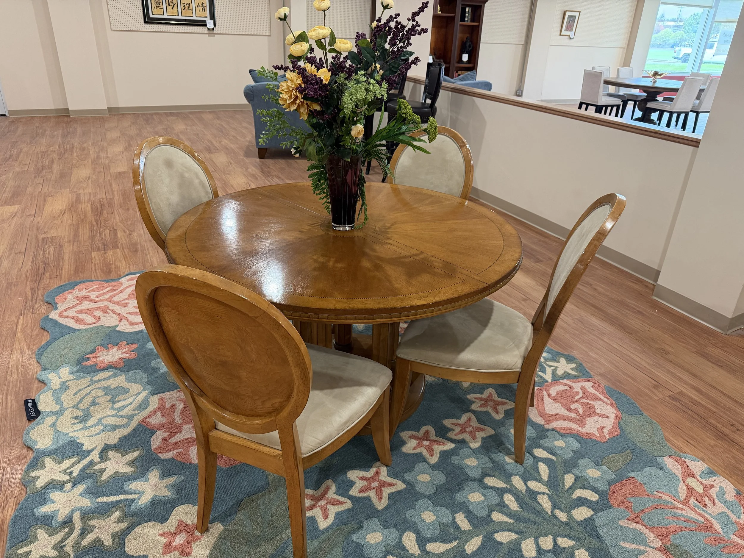 A wooden round dining table with a large floral arrangement in a tall, dark vase, placed on a colorful, floral patterned rug in a bright room with hardwood floors.