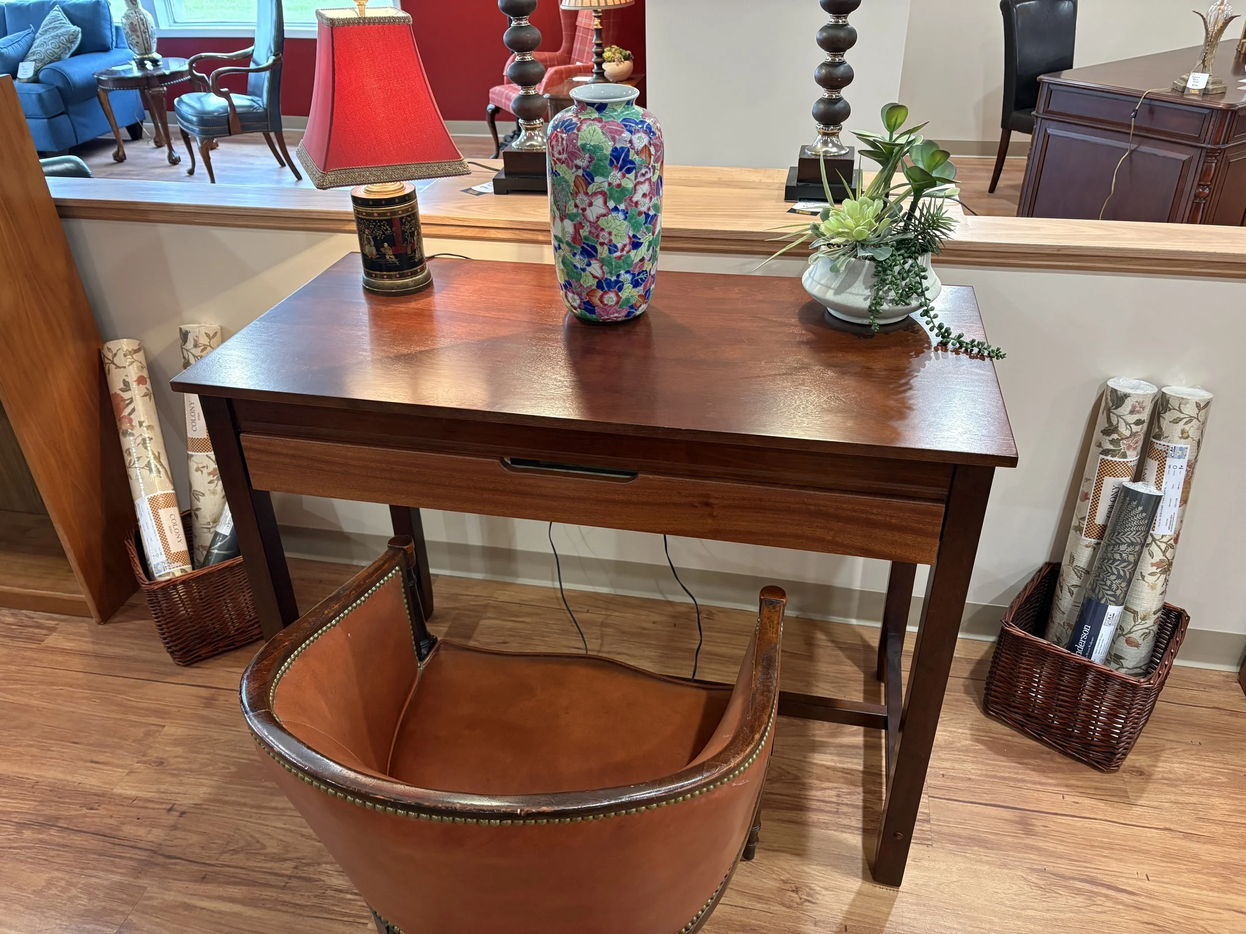 A wooden dining table with a decorative colorful floral vase, a table lamp with a red shade, and a potted green plant on top, surrounded by chairs in a furniture store with various other furniture and rolled wallpaper on the sides.