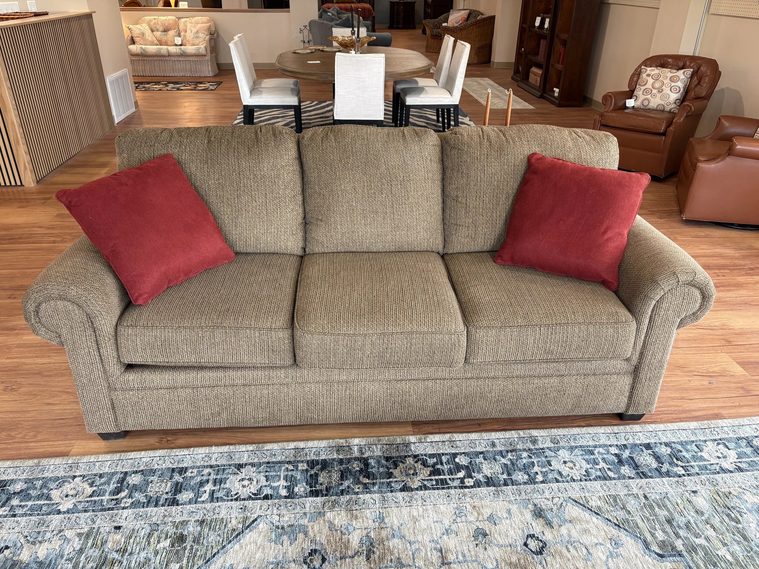 A living room storefront with a beige upholstered sofa in the foreground accented with red throw pillows. Behind the sofa, there is a dining table with six white chairs. To the right, there are two brown leather armchairs, and a plush ottoman. The ro