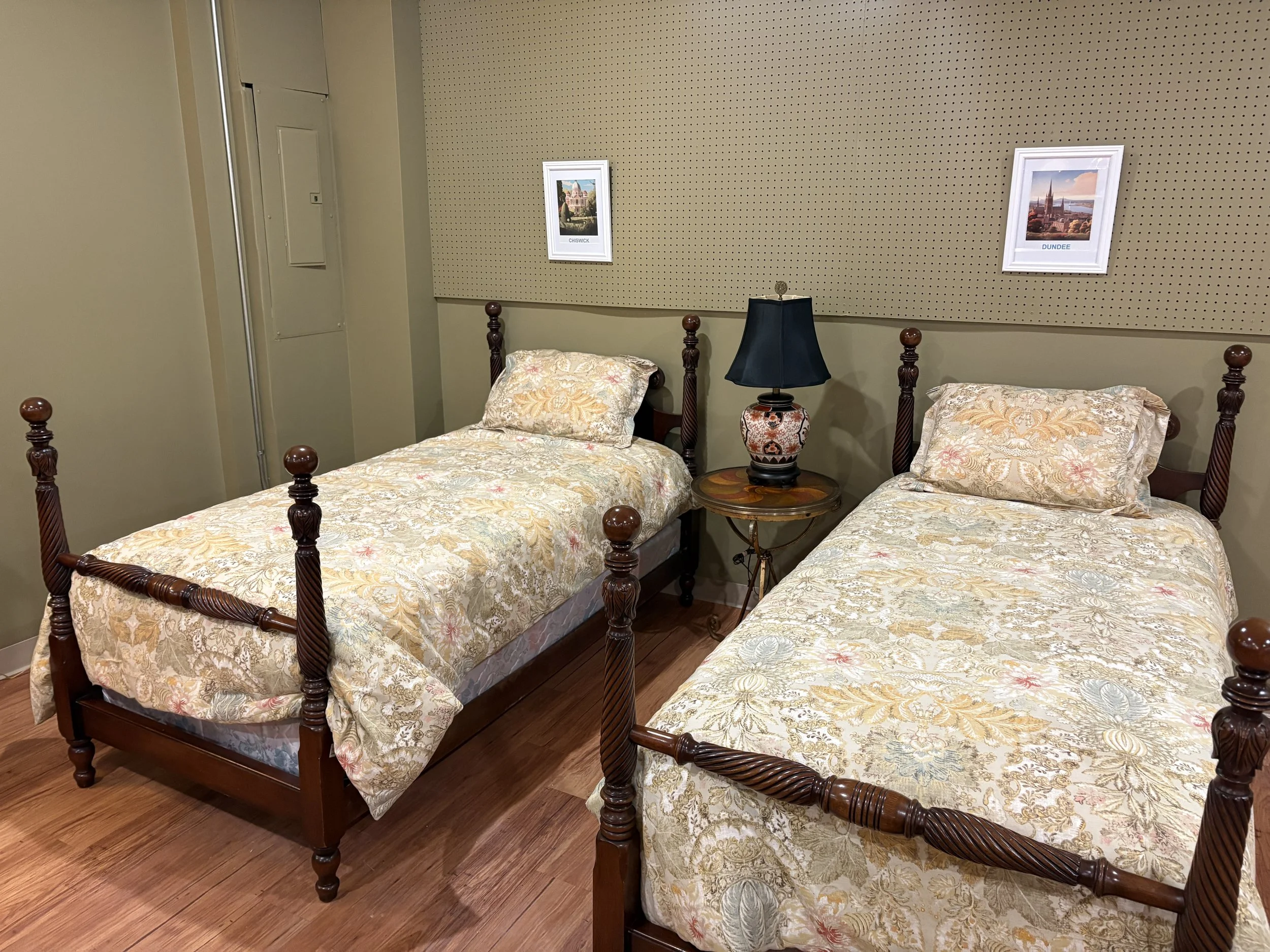 Two twin beds with floral bedding and carved wooden bed frames in a bedroom. A black lampshade on a small wooden side table is between the beds. Two framed pictures hang on the wall above the beds, one labeled 'Chiswick' and the other 'Dundee'.