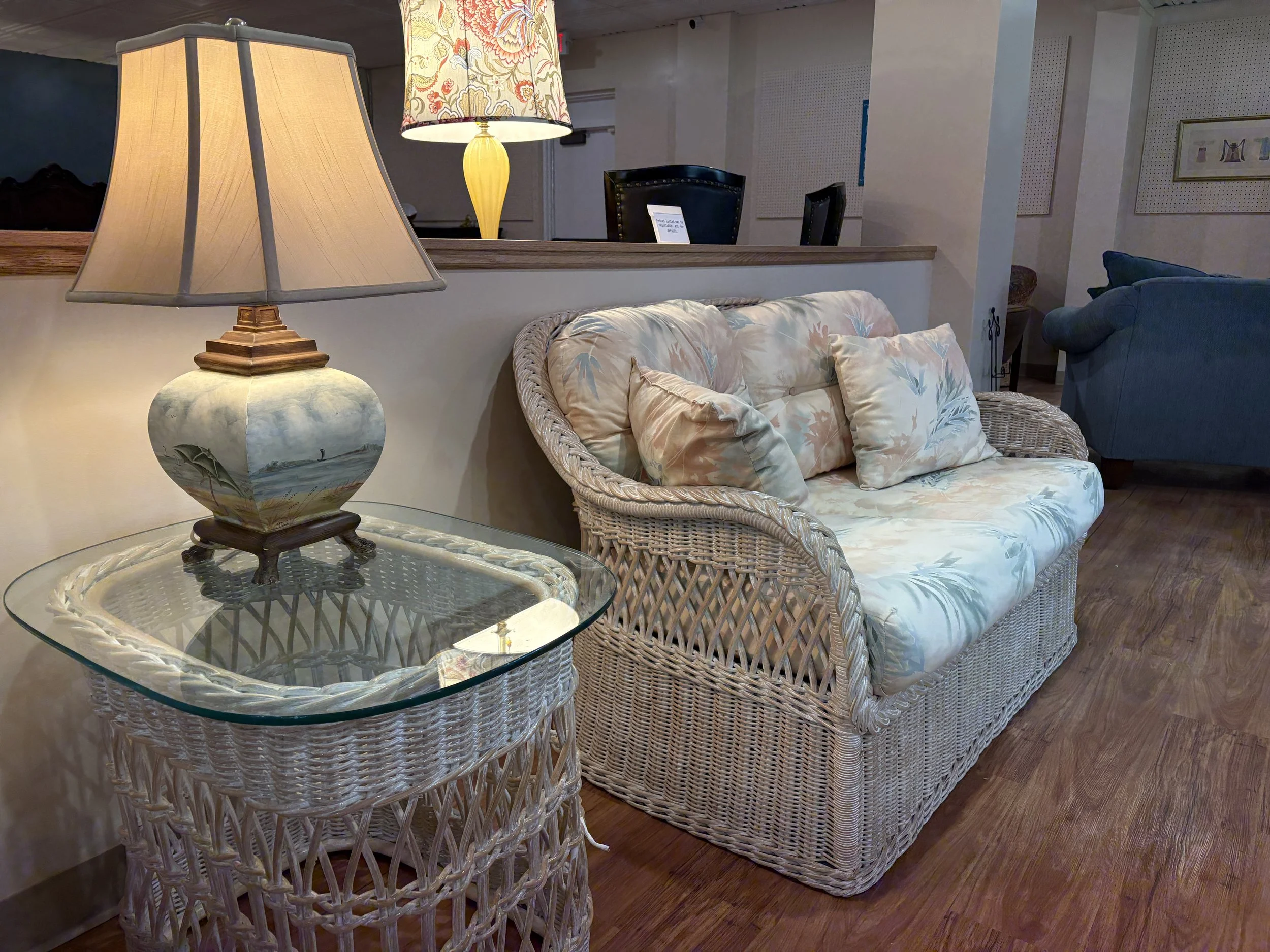 A cozy living room with a wicker sofa adorned with floral cushions, a glass-topped wicker coffee table, and a table lamp with a landscape painting on its base. In the background, there are additional chairs and framed pictures on the wall.
