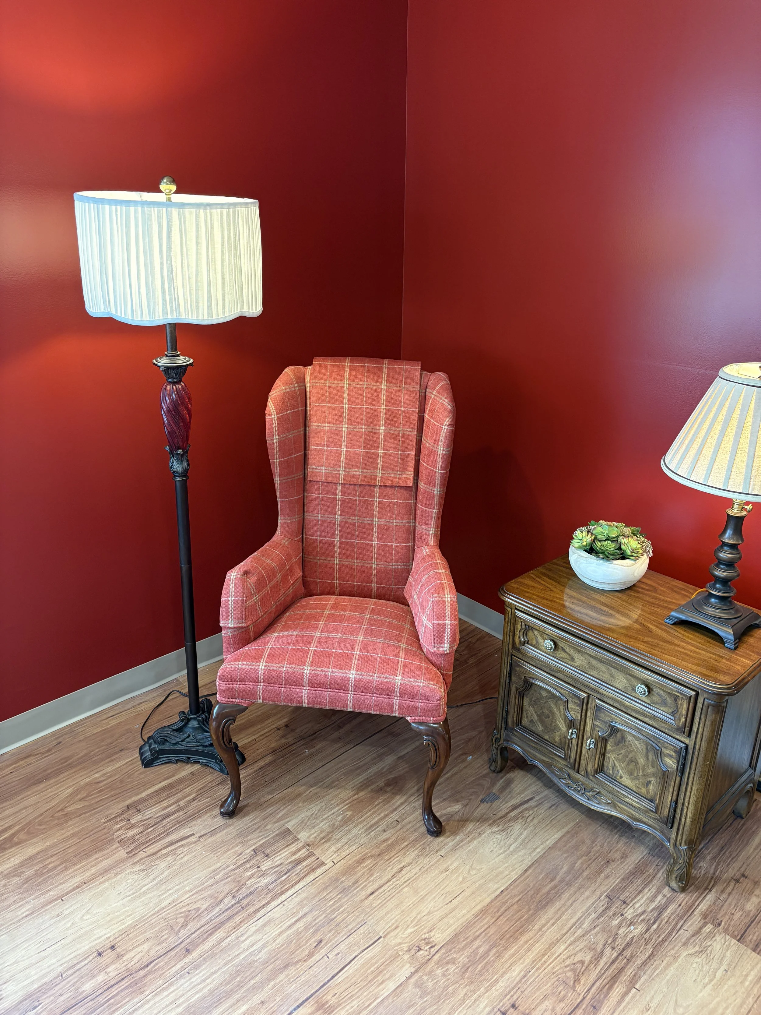 A cozy corner with a red plaid armchair, a vintage wooden side table with a drawer, a potted succulent plant, a tall floor lamp with a striped cream and blue lampshade, against a deep red wall and wooden plank flooring.