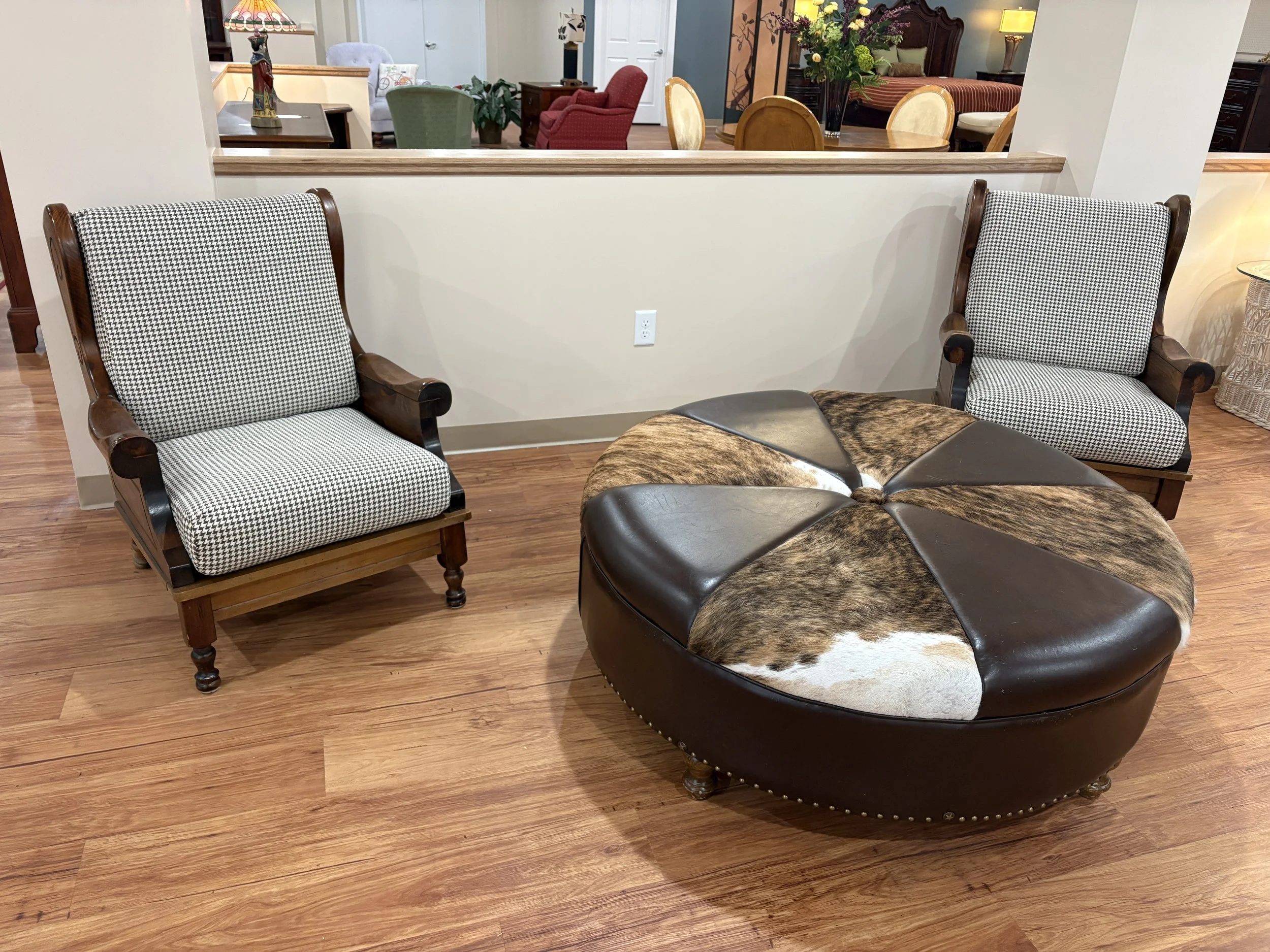 Living room with two black and white houndstooth upholstered wooden armchairs, a round leather ottoman with a cowhide print, and a beige wall with an electrical outlet. In the background, there is a dining area with a table and chairs, a flower vase,