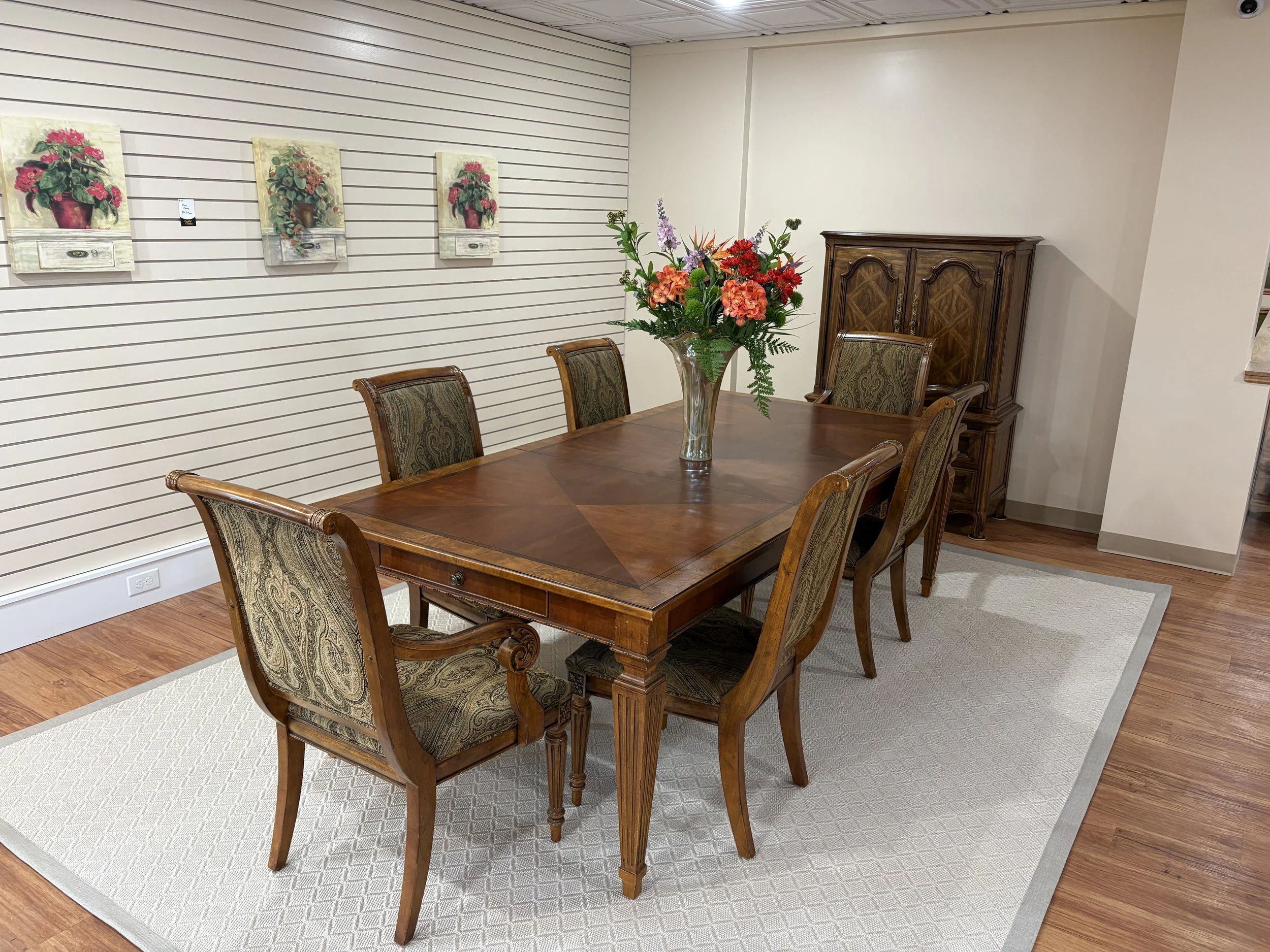 A wooden dining table with six upholstered chairs, a large glass vase with a colorful flower arrangement, a wooden china cabinet in the corner, white shiplap wall with three floral paintings framed in beige, beige walls, hardwood flooring, and a beig