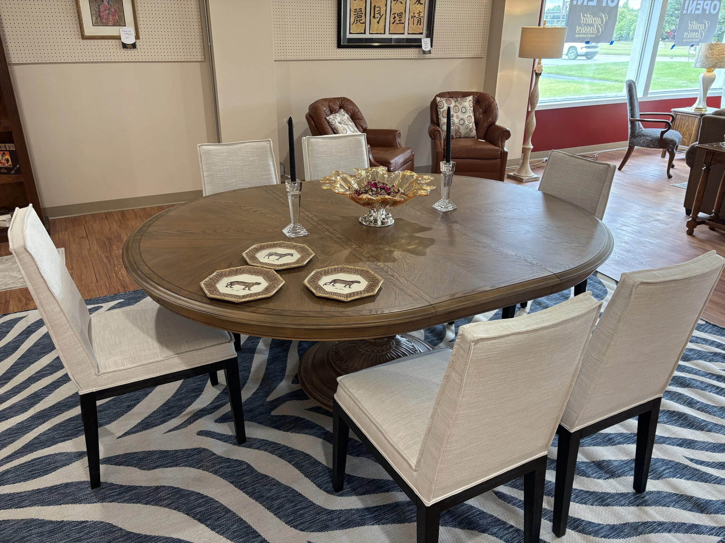 A dining room with a large wooden oval table surrounded by white upholstered chairs. On the table are decorative plates with horse illustrations and a gold-colored centerpiece bowl filled with dried flowers. In the background, there are two brown lea