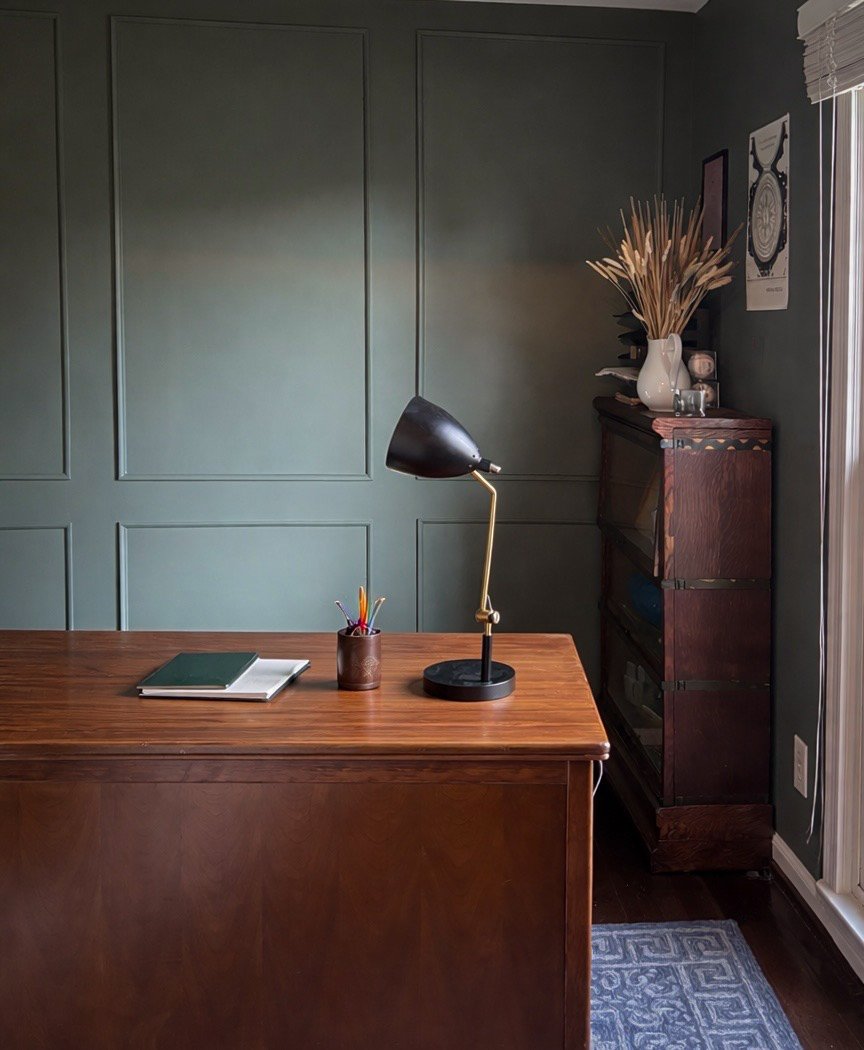 A minimalist desk setup featuring a wooden desk with a black and brass adjustable desk lamp, an open green notebook, and a container with colored pencils. There's a dark wooden cabinet on the right, topped with a white vase holding dried plants, behind which are framed artwork and a window with white blinds.