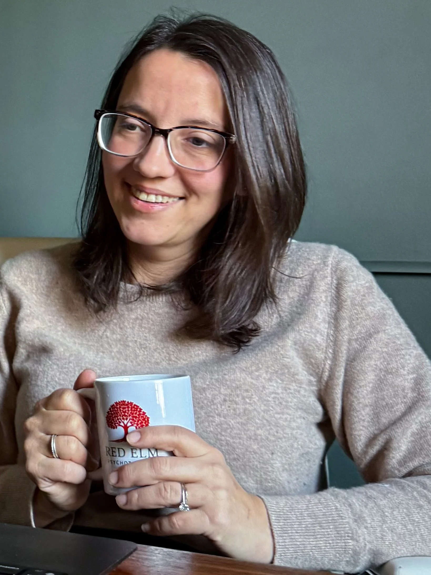 Woman with glasses smiling, holding a mug with a red tree logo, sitting at a desk.