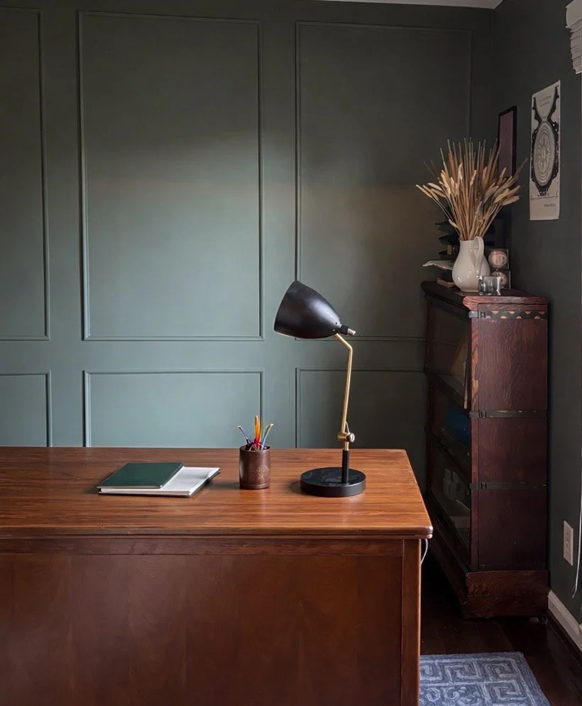 A home office with a wooden desk, a black and gold desk lamp, a notebook and a pen holder on the desk, a dark wooden bookshelf in the corner with decorative items, a white vase with dried plants, and wall art, against a green paneled wall.